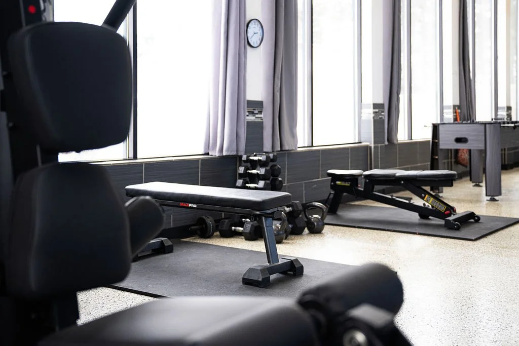 Empty gym workout area with adjustable benches, dumbbells, and a foosball table near large windows with curtains.