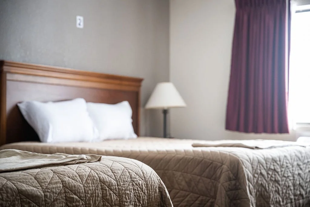 Resident room with two beige beds, white pillows, a beige quilted bedspread, a wooden headboard, a white table lamp, and purple curtains covering a window.