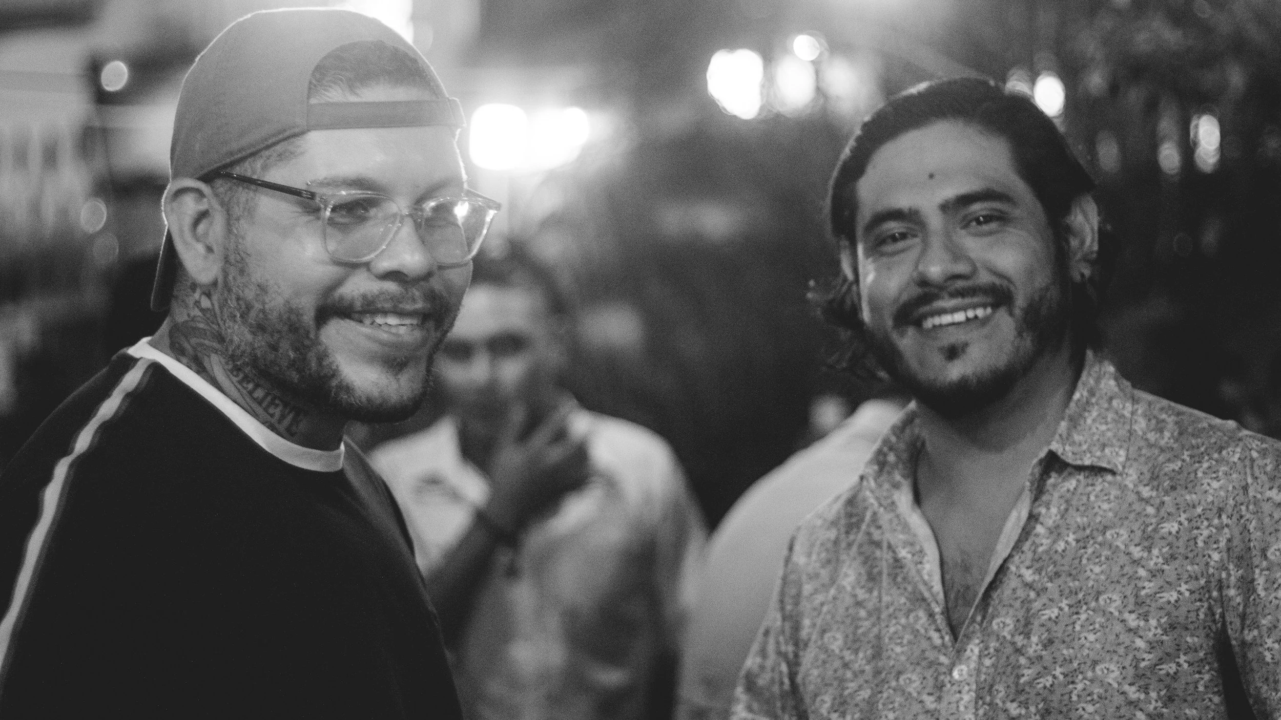 Two smiling men at night, one with glasses and a baseball cap, and the other with long hair and a patterned shirt, standing outdoors with blurred lights in the background.