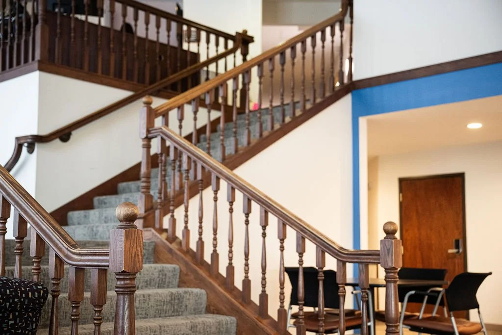 Wooden staircase with gray carpeted steps and wooden balustrades in a building interior with white and blue walls and a small seating area.