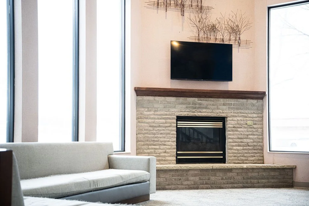 Living room with beige sofa, stone fireplace, flat-screen TV above the mantle, and large windows bringing in natural light.