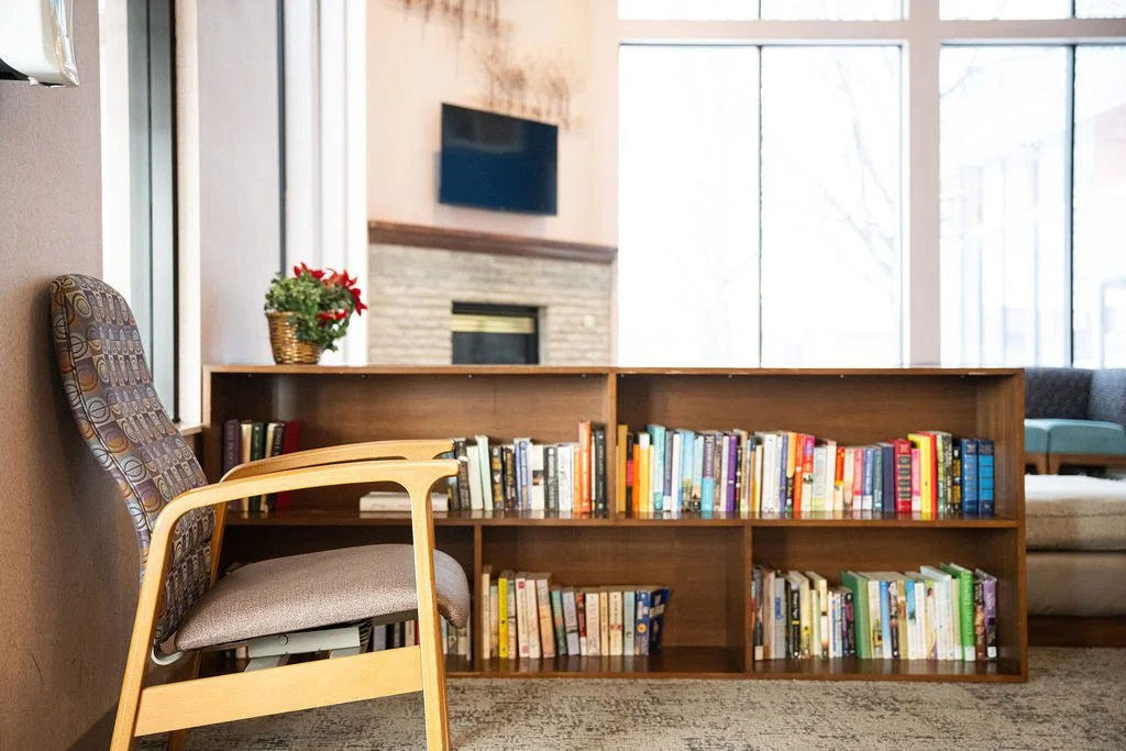 A cozy indoor reading area with a wooden bookshelf filled with books, a padded armchair, and a potted plant with red flowers, near a window with large glass panes.