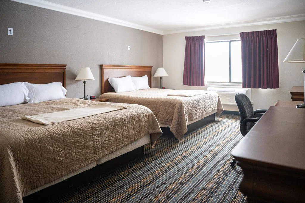 Resident room with two beds, nightstands, lamps, window with red curtains, desk with chair, and carpeted floor.