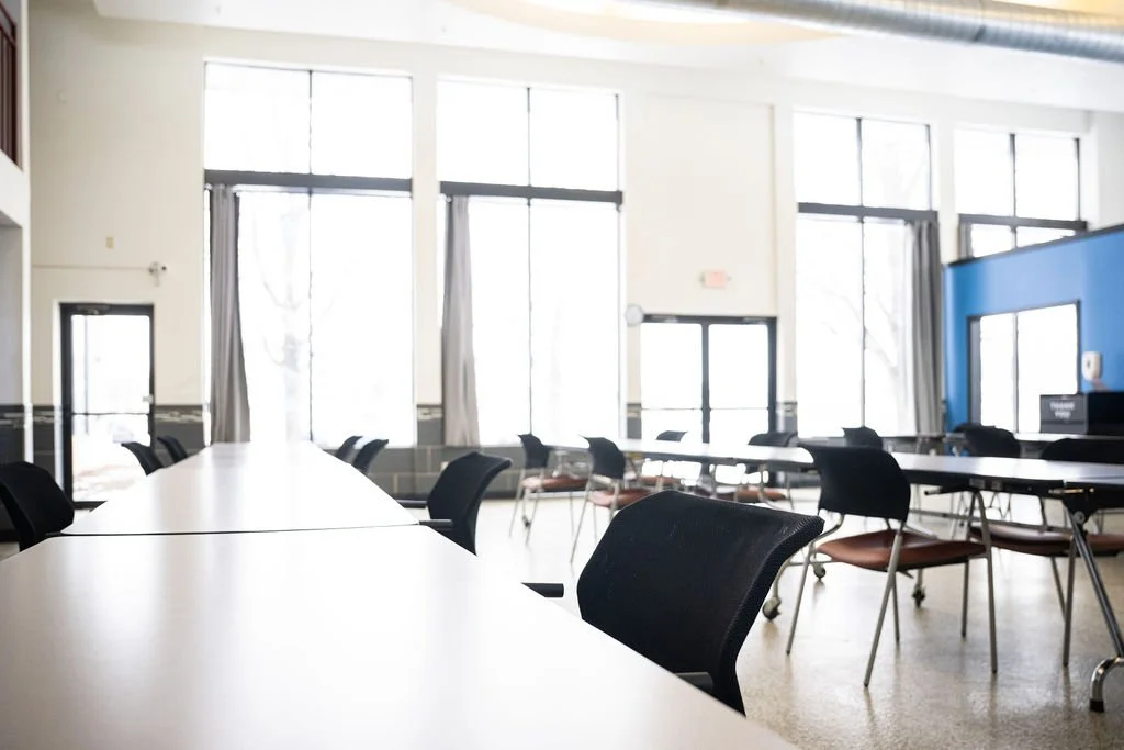 Empty gathering and eating room with long table, black chairs, large windows with curtains, and a blue accent wall.