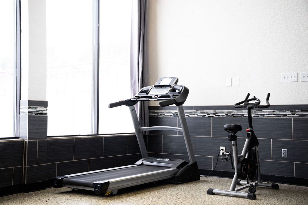 image showing a treadmill and a stationary bike in a modern gym with large windows and white walls.