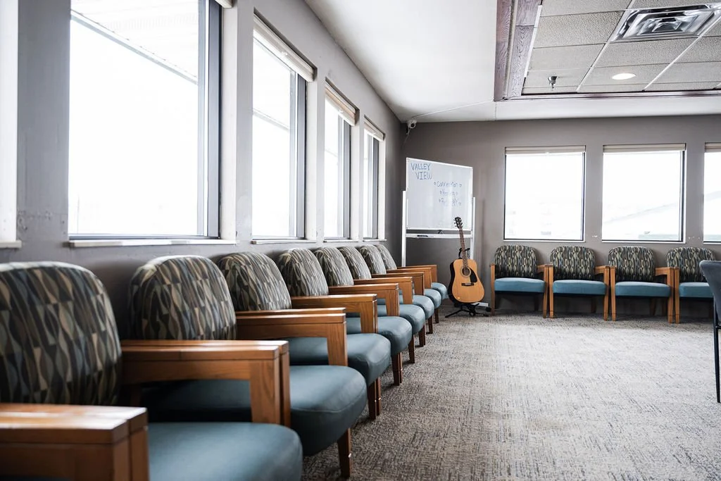 Empty waiting room with row of patterned chairs, a guitar, and a whiteboard in a building with large windows.