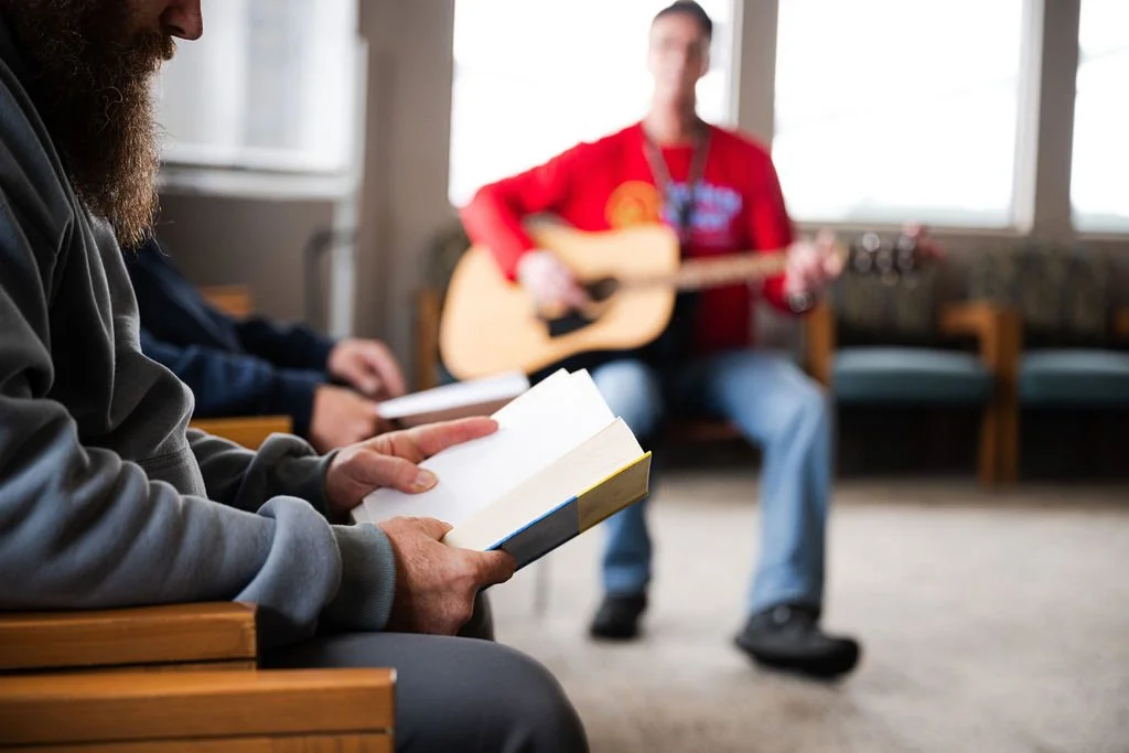 Person reading a book in the foreground with a musician playing guitar in the background during a group gathering.