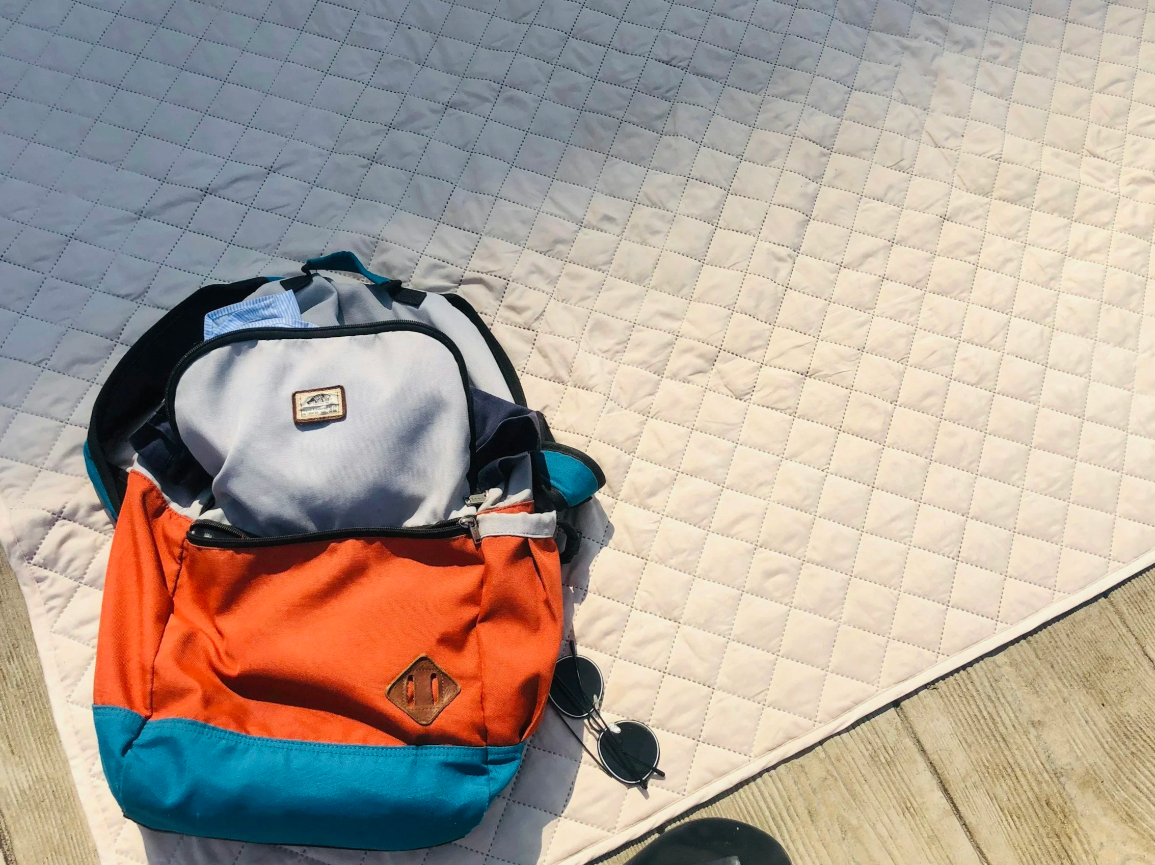 Colorful backpack and sunglasses on a white quilted mattress on a beige floor.