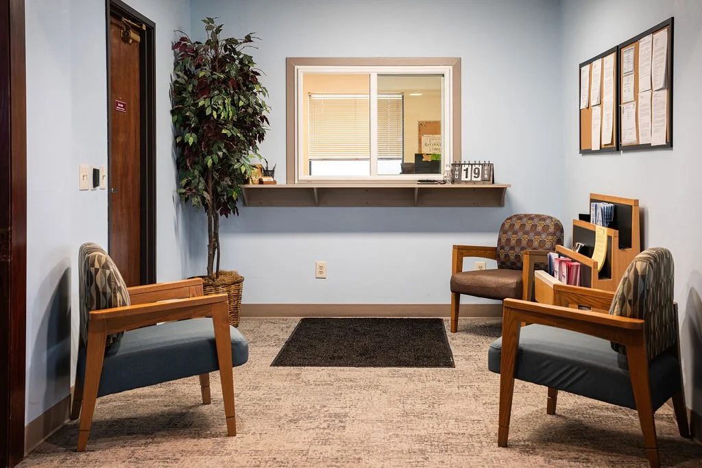 A small waiting room with two chairs with blue cushions and wood armrests, a third chair with a patterned fabric cushion, a tall potted plant, and a desk with files and a book organizer. There's a window with blinds, a bulletin board on the wall, and