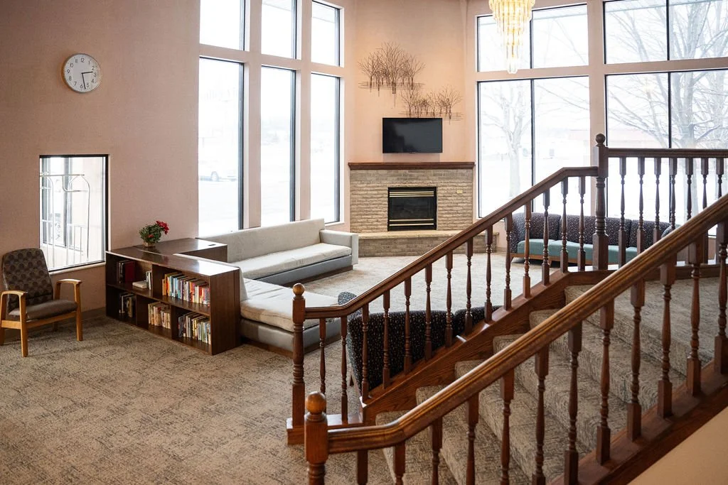 A spacious living room with large windows, a stone fireplace with a flat-screen TV above, a beige sofa, a wooden bookshelf with books, and a staircase with wooden railing leading upstairs.