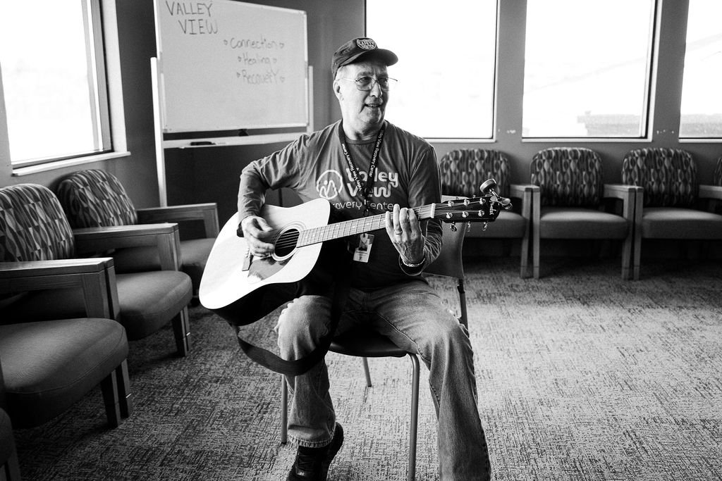 A man wearing a cap and lanyard sitting on a chair, playing an acoustic guitar in a room with multiple chairs and windows at Valley View Recovery Center