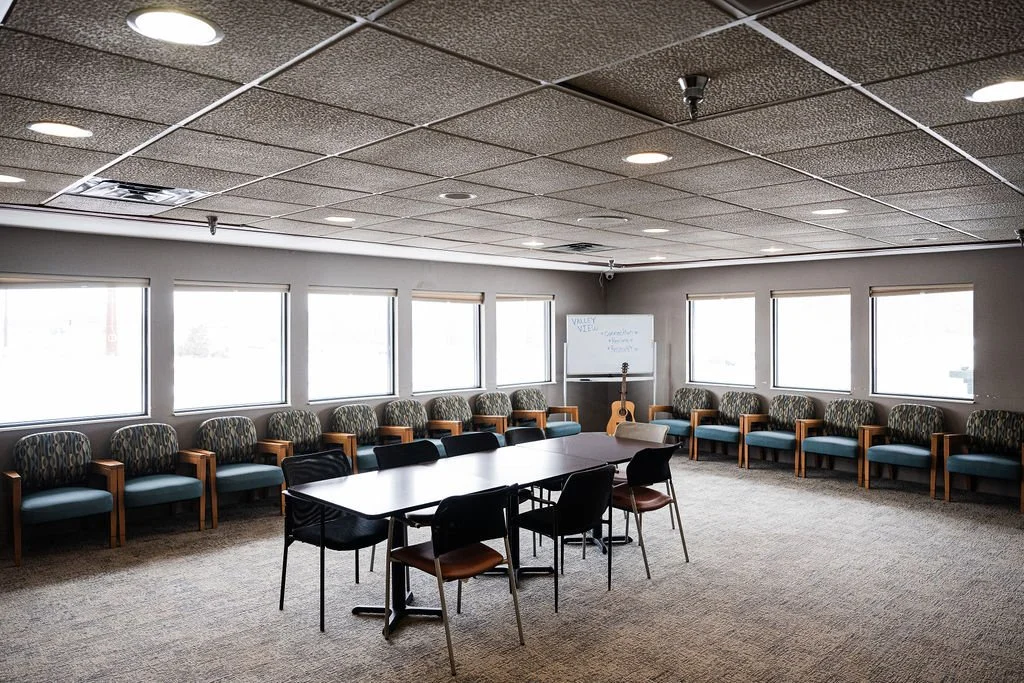 Empty common space room with a large table and chairs, a whiteboard, and a guitar, surrounded by windows.