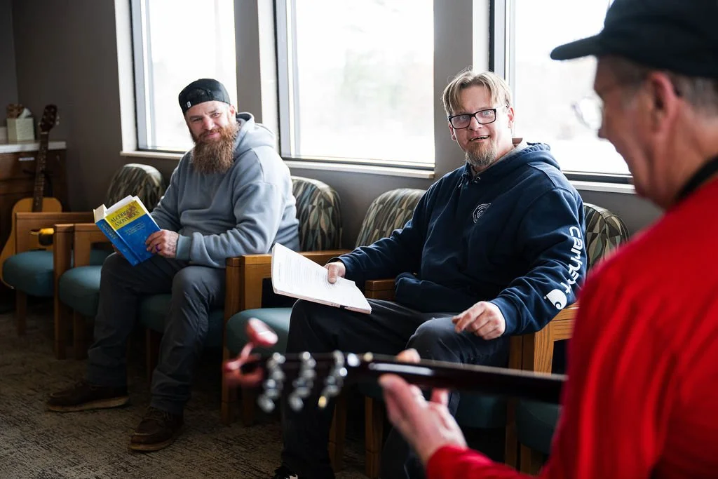 Three men sitting and socializing in a room with large windows. One man is playing guitar, another is holding a booklet, and the third man is looking at the guitarist, smiling.