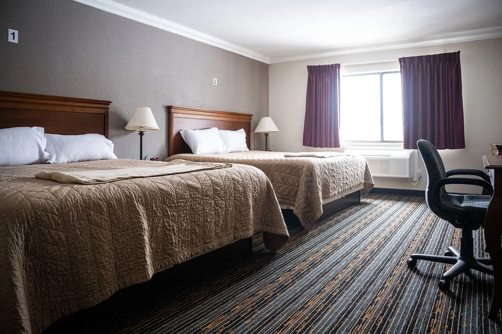 Two double beds with wooden headboards, beige bedding, and white pillows in a room at Valley View Recovery Center with a window with purple curtains, a desk, and a black chair.