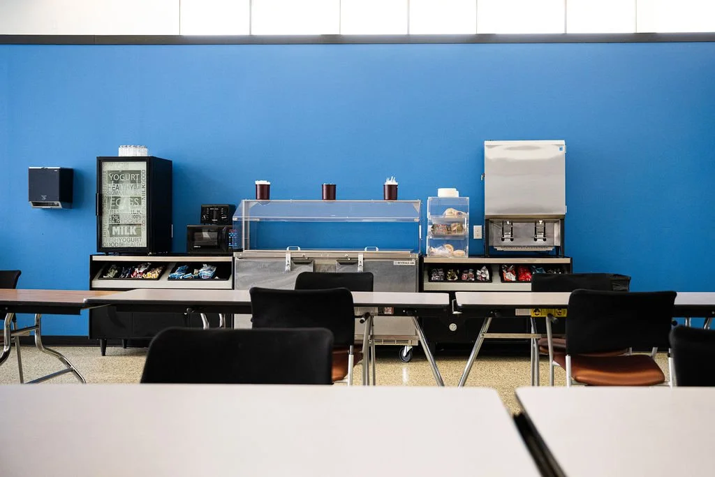 Empty snack and drink station with a blue wall behind it, containing a vending machine, a microwave, a glass display case with beverages, and snack shelves, with chairs and tables in the foreground.