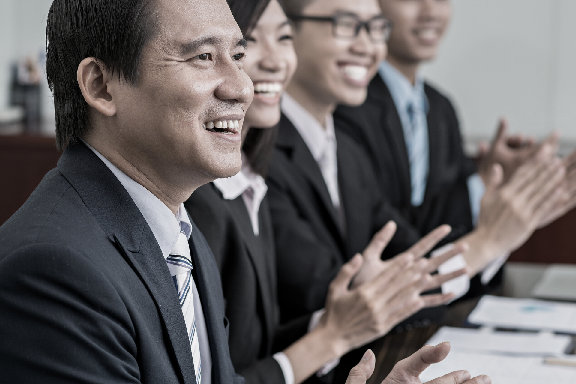 A group of professional businesspeople sitting at a conference table, smiling and clapping during a meeting.