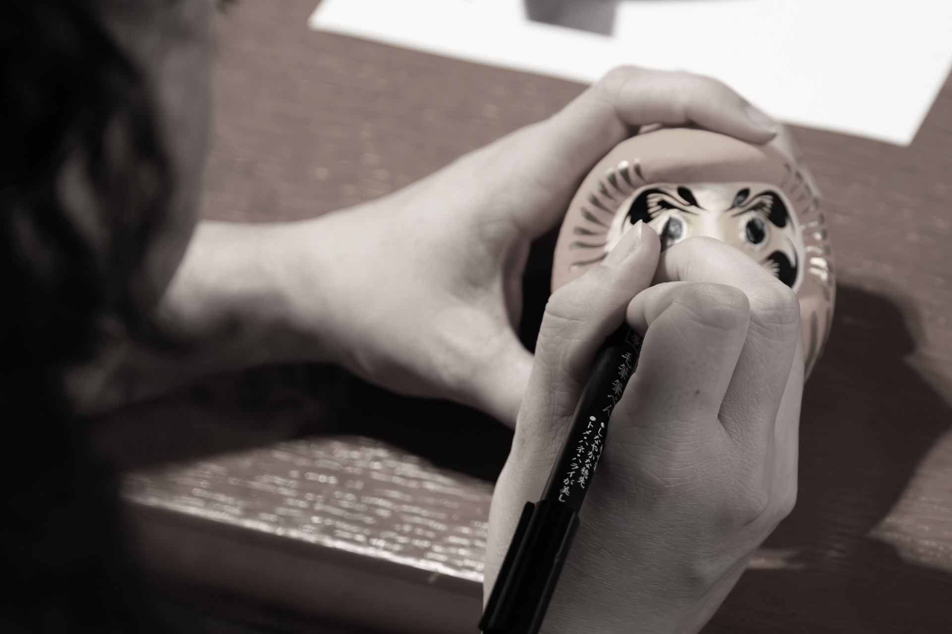 A person is drawing on a Daruma doll with a black marker, focusing on the doll's face, placed on a wooden surface with papers nearby.