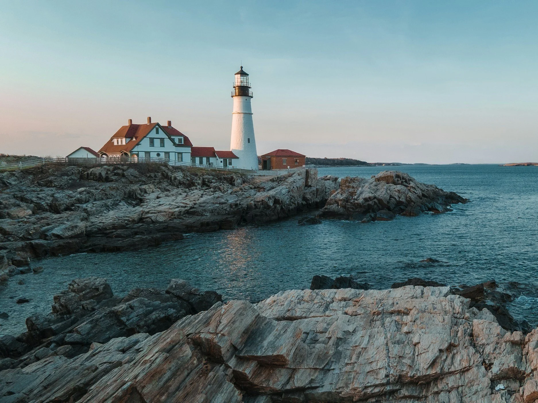 A lighthouse standing on a rocky shoreline with a house nearby, overlooking the ocean at sunset.