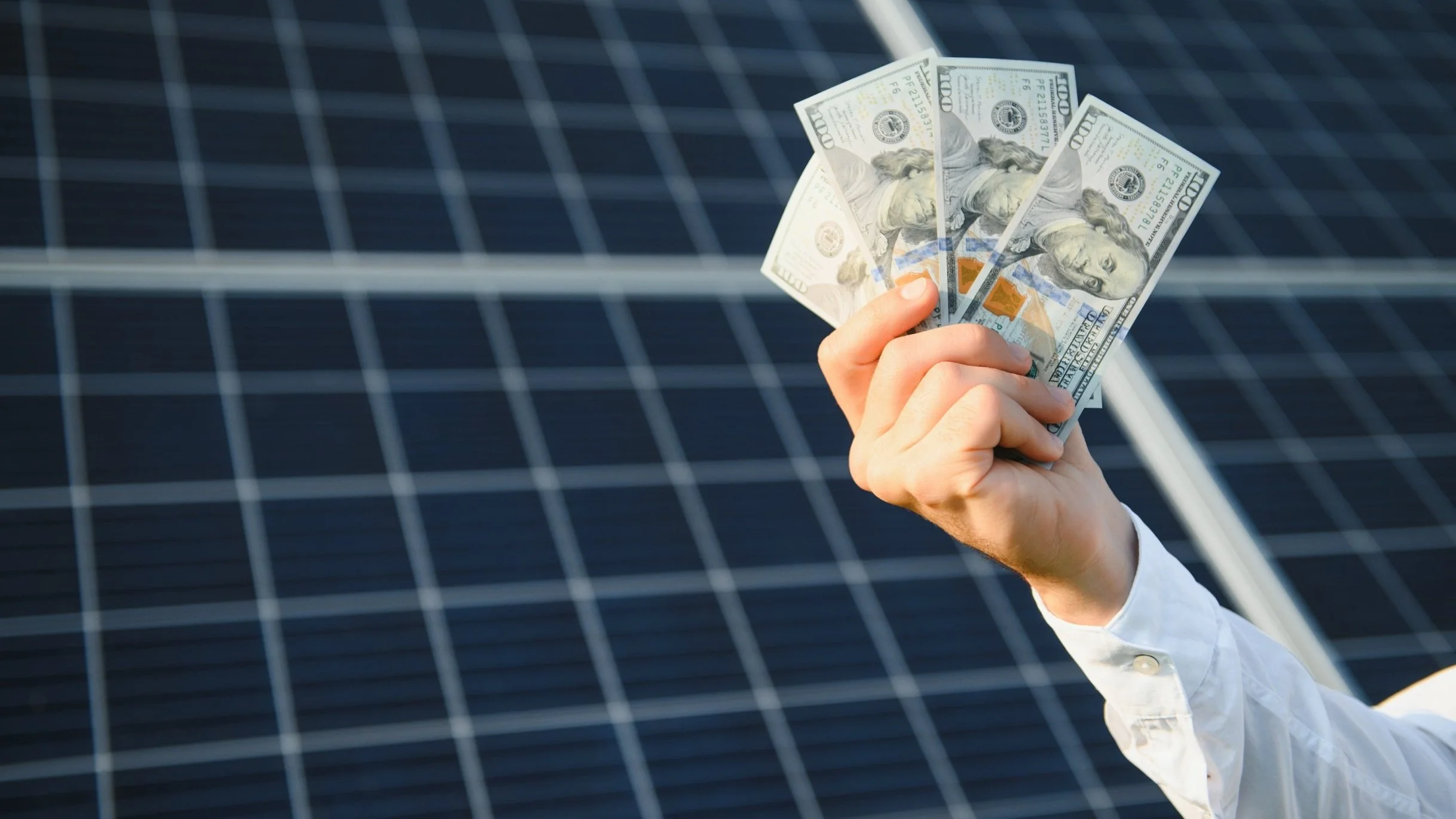 A hand holding three hundred dollar bills in front of solar panels.