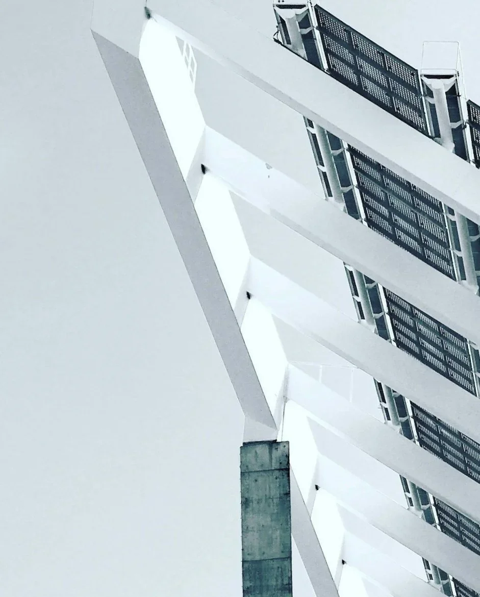 Modern architectural structure with a large, overhanging roof and solar panels, supported by tall concrete pillars, with two small people walking beneath it.