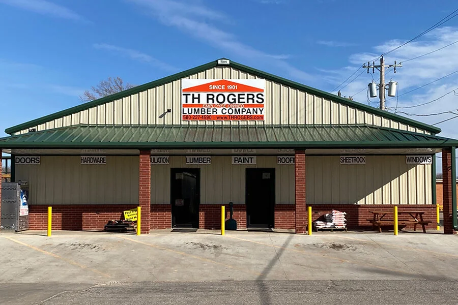 Front view of The Rogers Lumber Company store with a sign indicating it sells doors, hardware, insulation, lumber, paint, roofing, sheetrock, and windows, and features a parking lot with yellow safety poles.