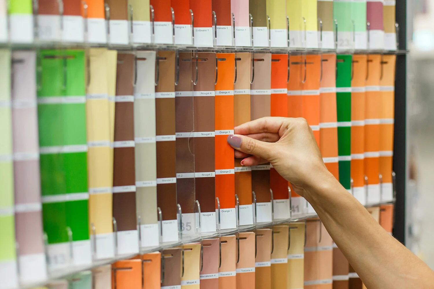 Person selecting a bright orange paint color from a wall of multiple paint swatches in a hardware store.