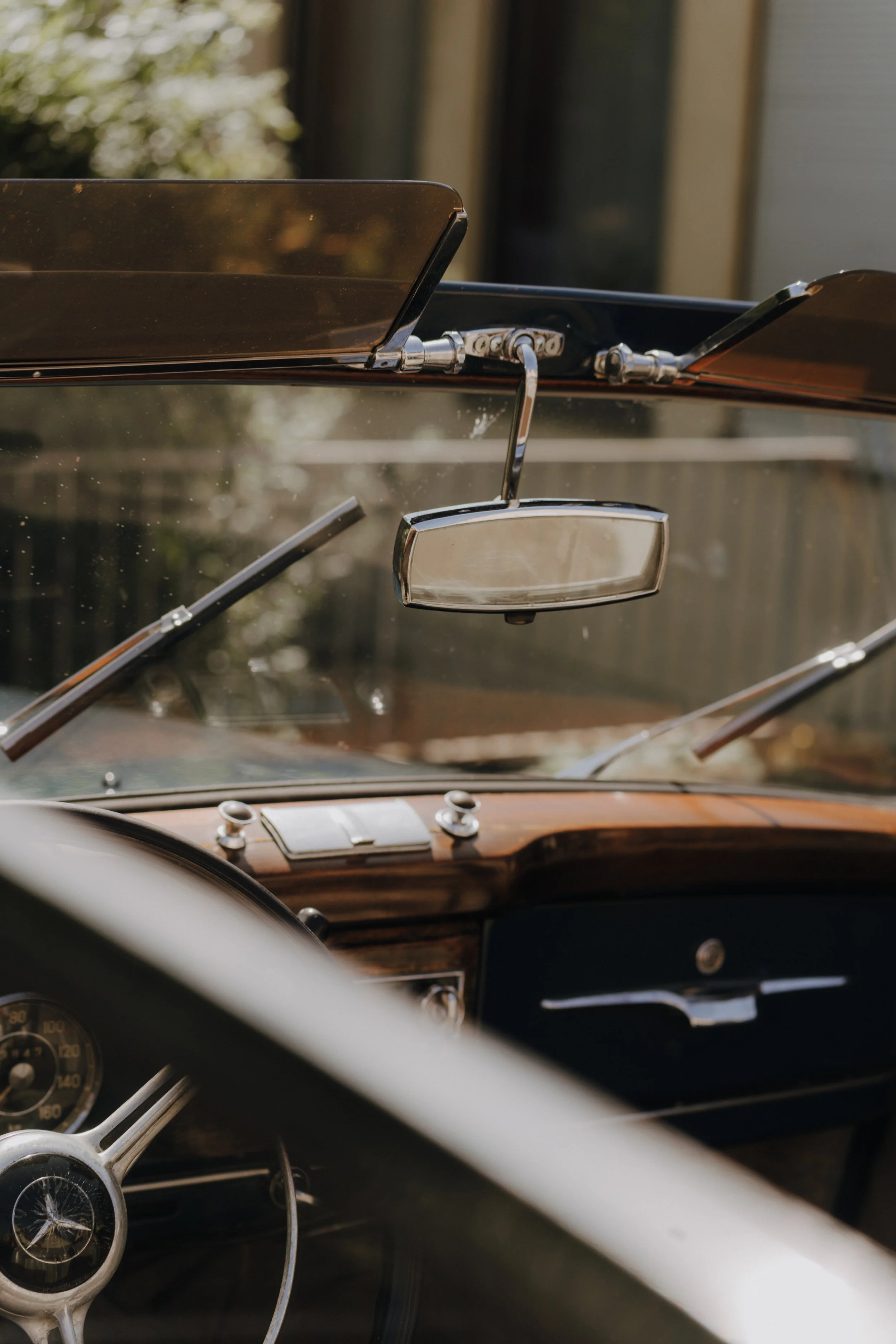 Innenraum eines Oldtimers mit Holzlenkrad, Spiegel und Armaturen. Blick durch die Windschutzscheibe nach draußen.