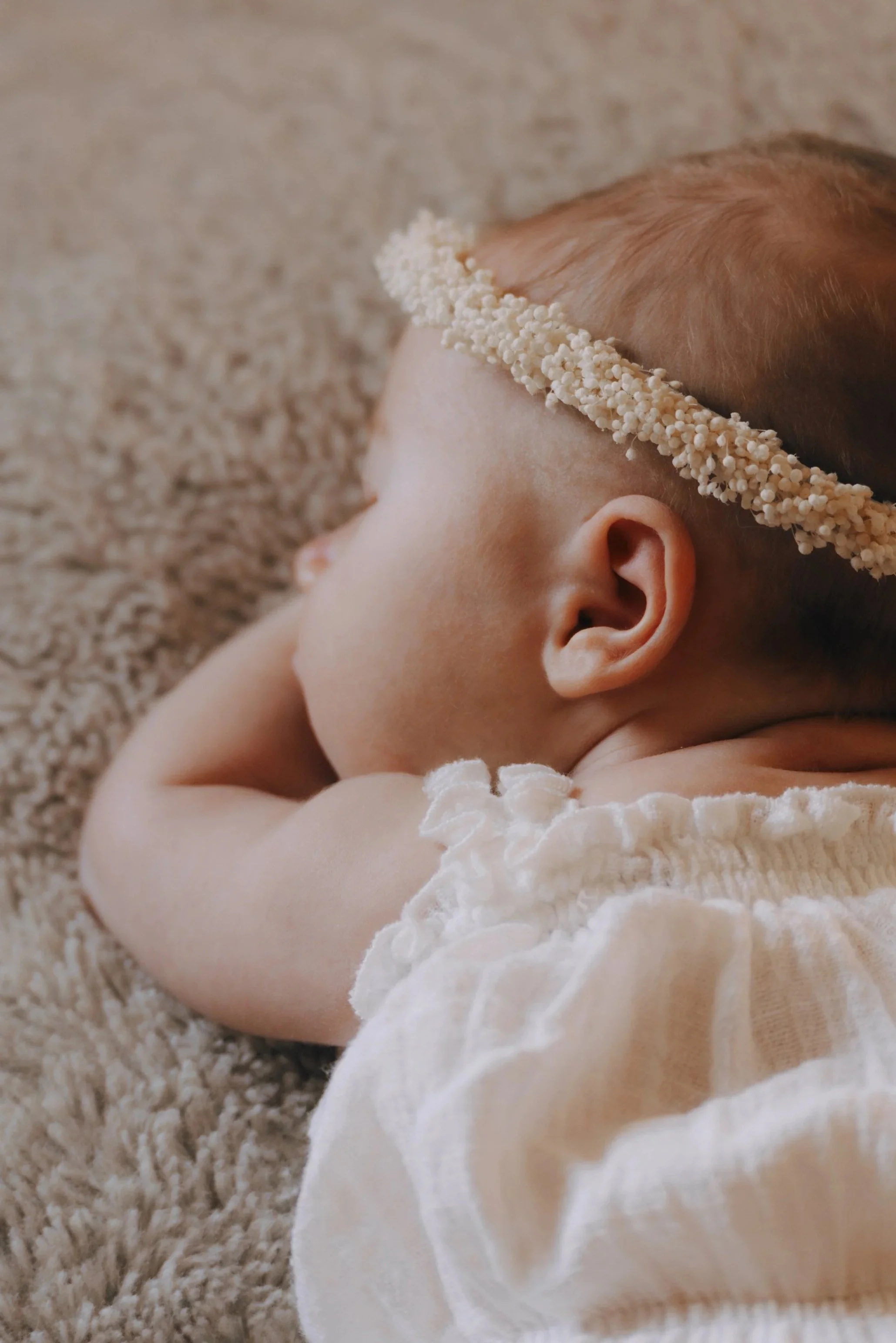 Nahaufnahme eines schlafenden Babys mit einem Blumenkranz im Haar auf einem weichen, pelzigen Untergrund.