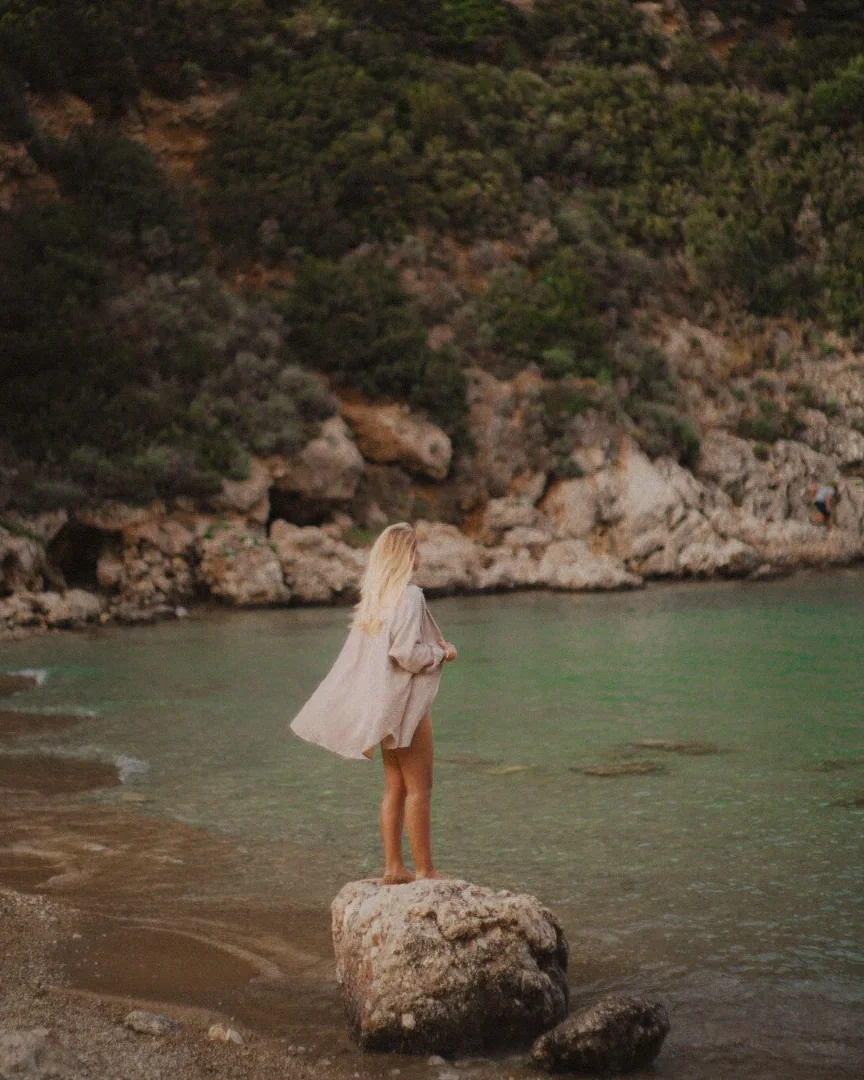 Frau steht auf einem großen Felsen am Strand, Blick auf das Meer, mit einer Küstenklippe im Hintergrund.