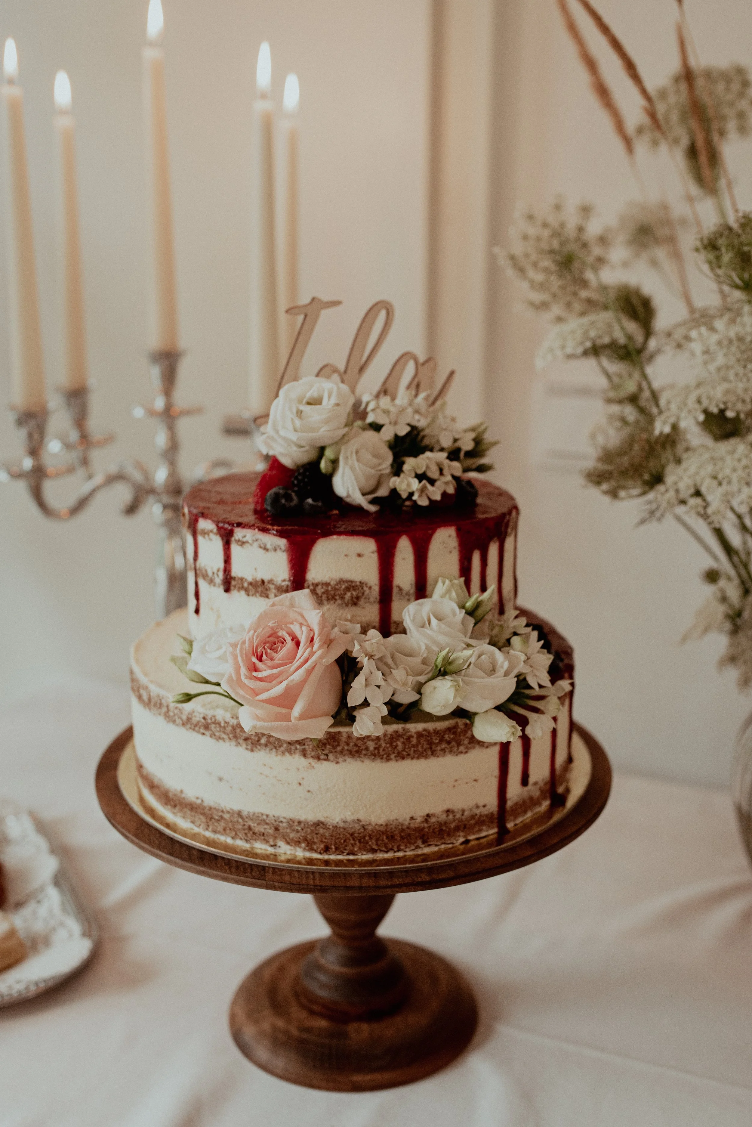 Hochzeitstorte mit Blumen und rotem Zuckerguss auf Holzständer, Kerzenleuchter im Hintergrund.