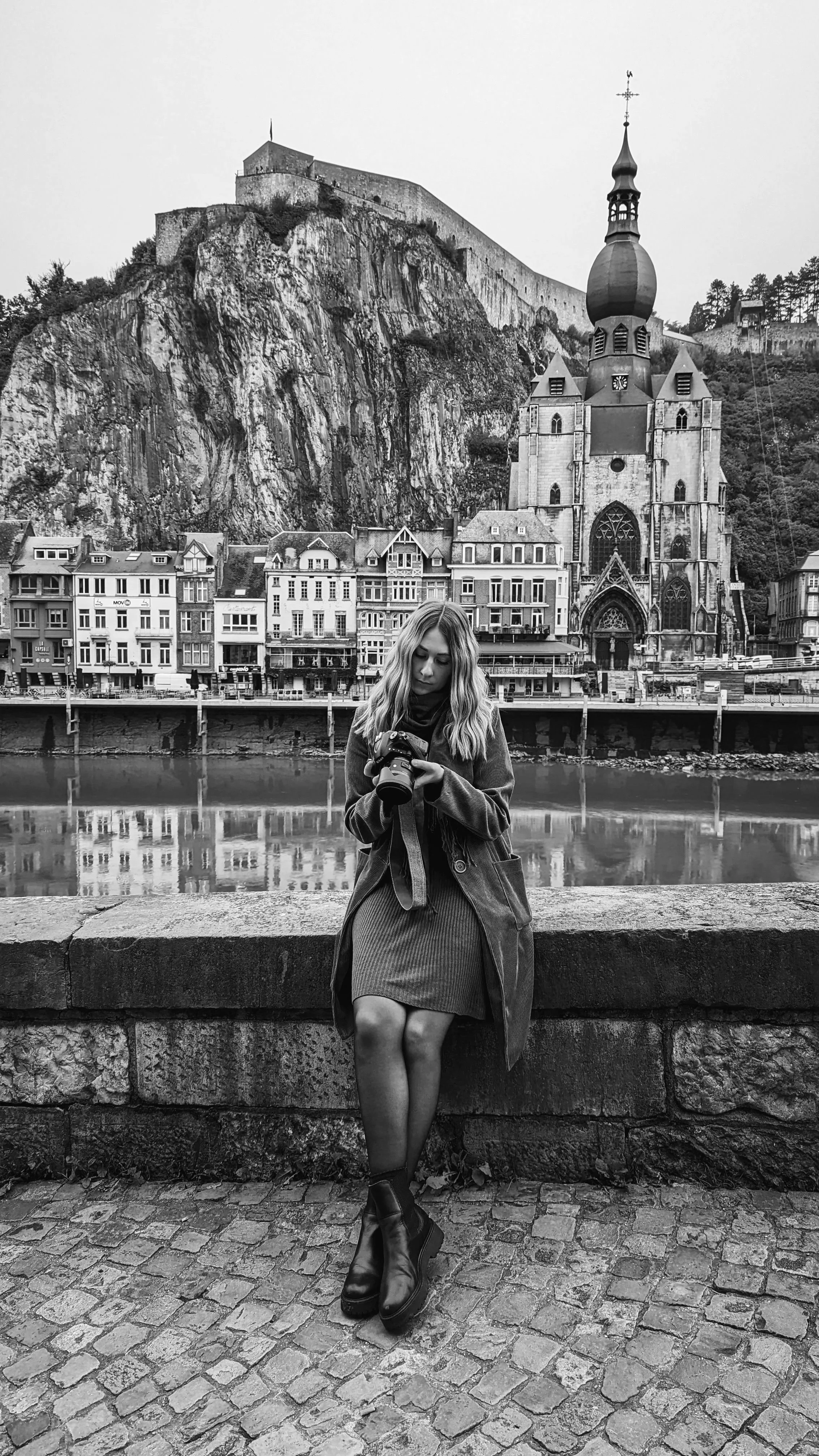 Eine junge Frau mit langen Haaren, trug einen Mantel, ein Kleid, Stiefel, sitzt an der Uferpromenade und schaut auf eine Kamera in ihren Händen. Im Hintergrund sind alte Häuser, eine Kirche mit Zwiebelkuppel und eine steile felsige Anhöhe mit einer Festung zu sehen.