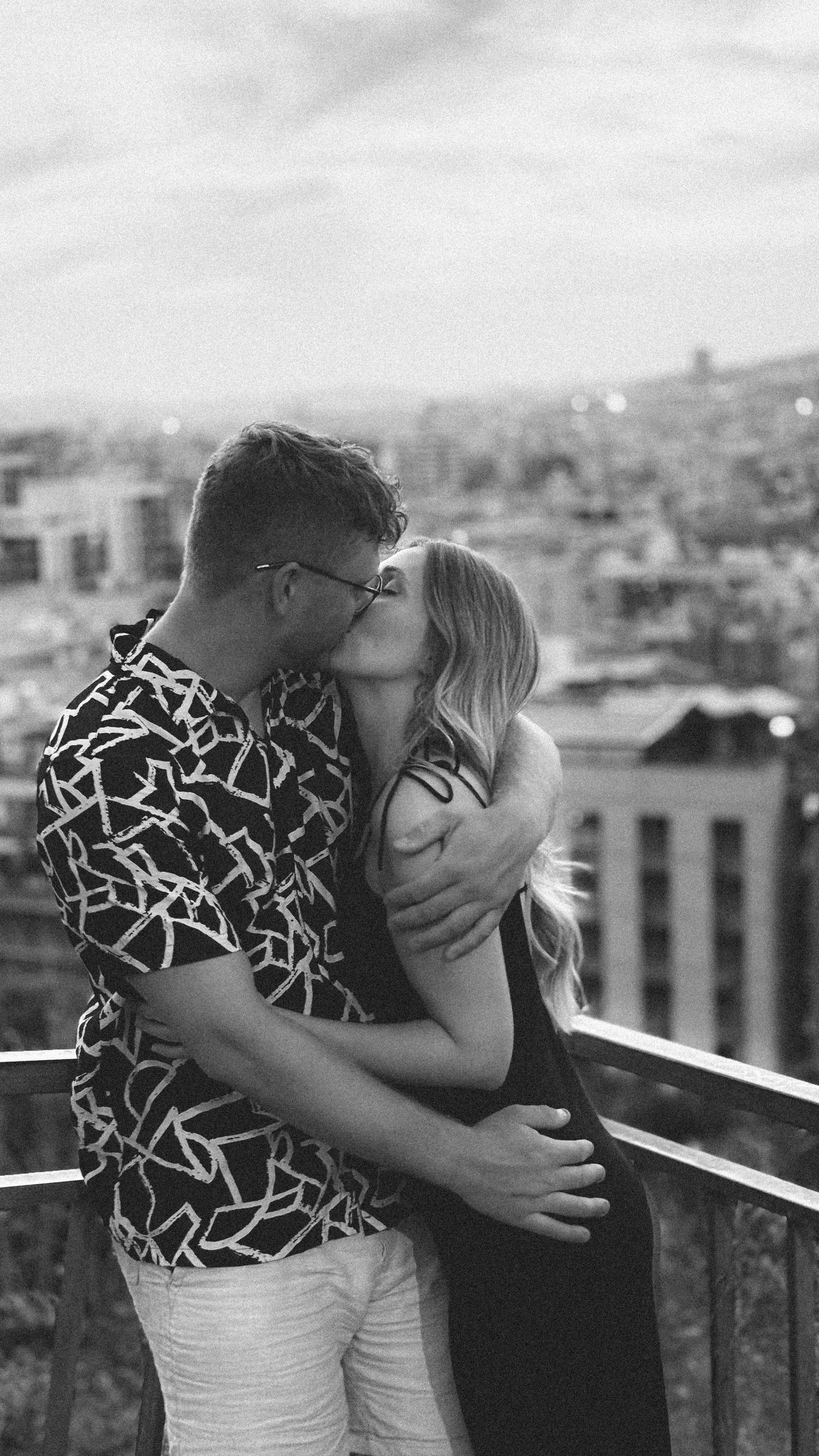 Ein verliebtes Paar küsst sich auf einem Balkon mit Stadtblick bei Sonnenuntergang, Schwarz-Weiß-Foto.