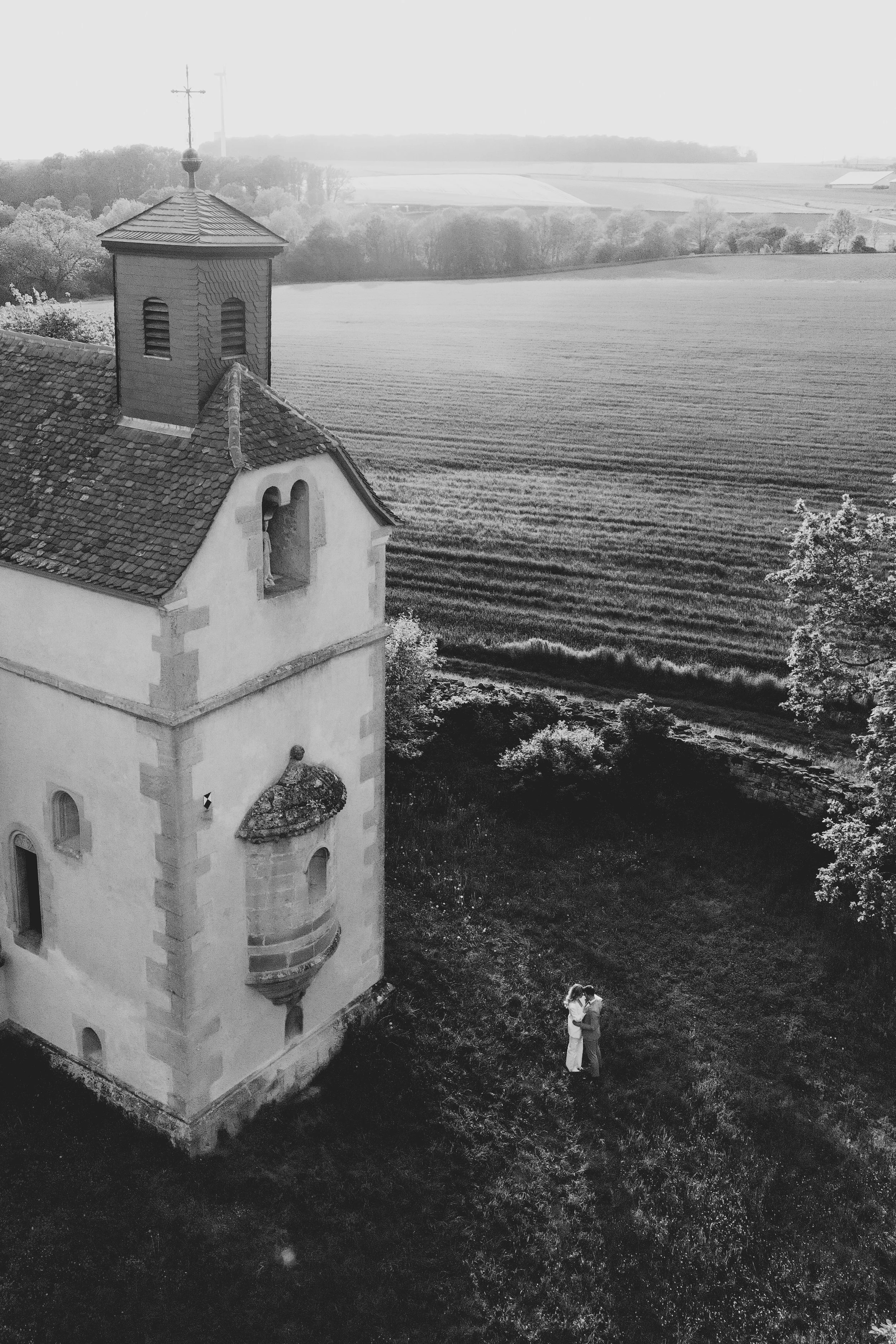 Ein schwarzes und weißes Foto einer Kirche mit einem Paar, das sich umarmt, im Vordergrund. Im Hintergrund weite Felder und Bäume unter einem bewölkten Himmel.