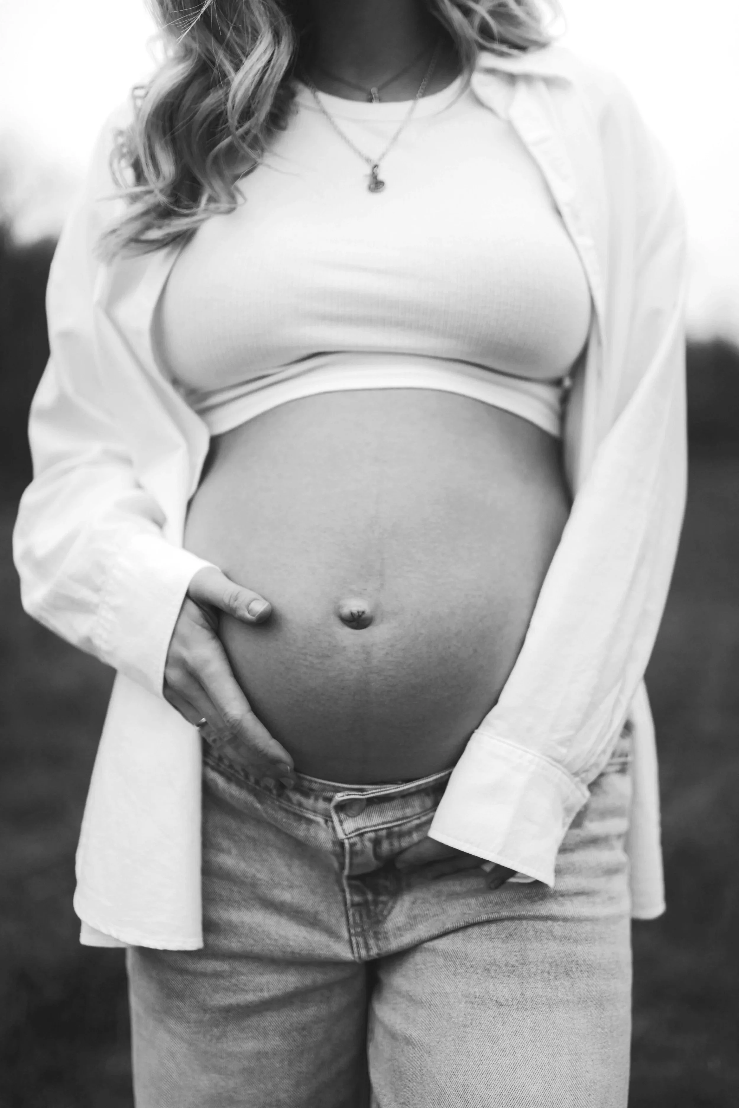 Schwangere Frau im Freien trägt ein weißes T-Shirt und eine offene Jacke, zeigt ihren Babybauch, trägt zwei Halsketten, einer mit einem kleinen Anhänger, und hat lockige Haare.