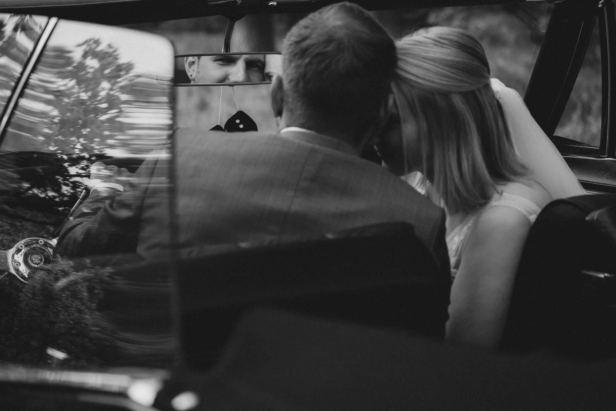 Ein Brautpaar sitzt in einem Auto, teilt einen Kuss, während sie sich im Rückspiegel ansehen, im Schwarz-Weiß-Stil.