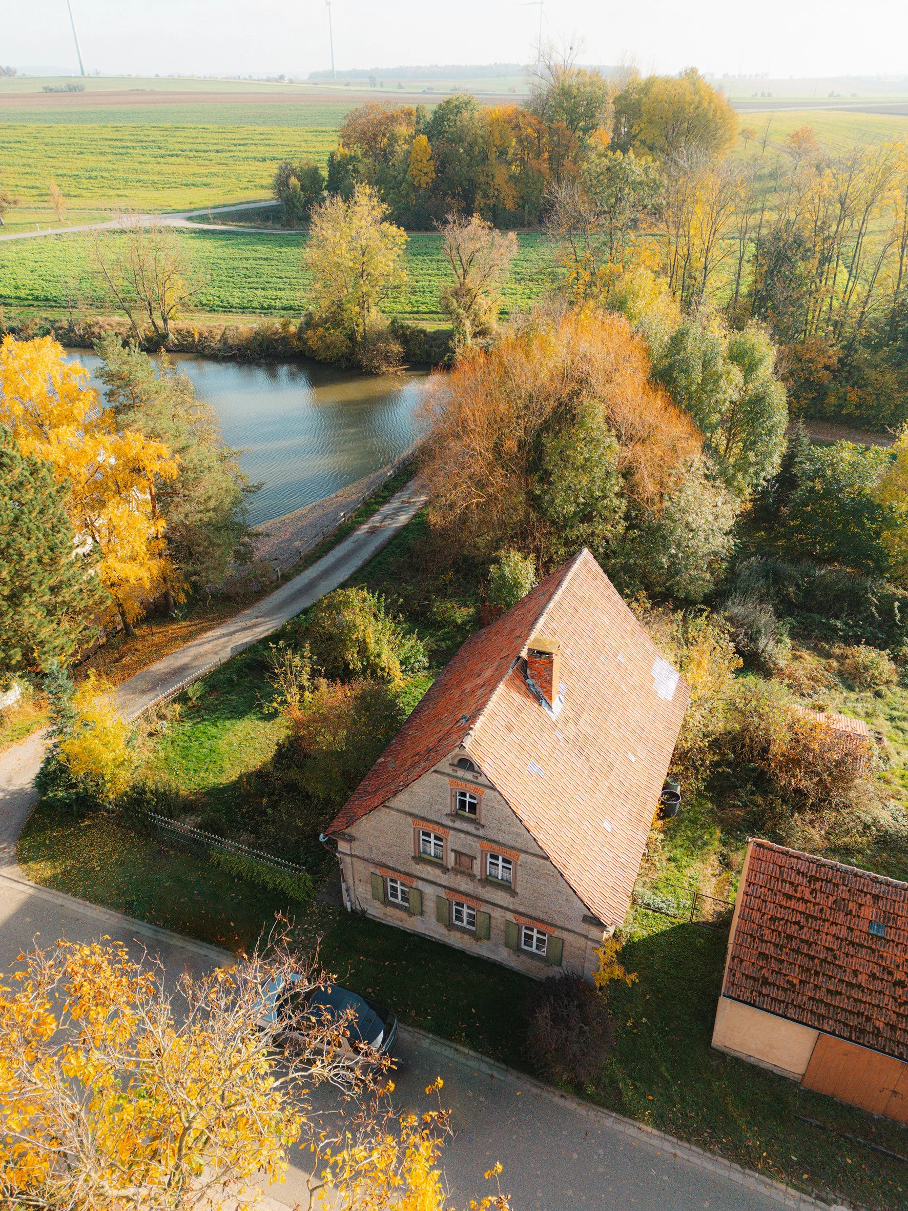 Vogelperspektive auf ein Haus mit rotem Dach, umgeben von Bäumen mit Herbstlaub, eine Straße, einen See und landwirtschaftliche Flächen im Hintergrund, bei sonnigem Wetter.