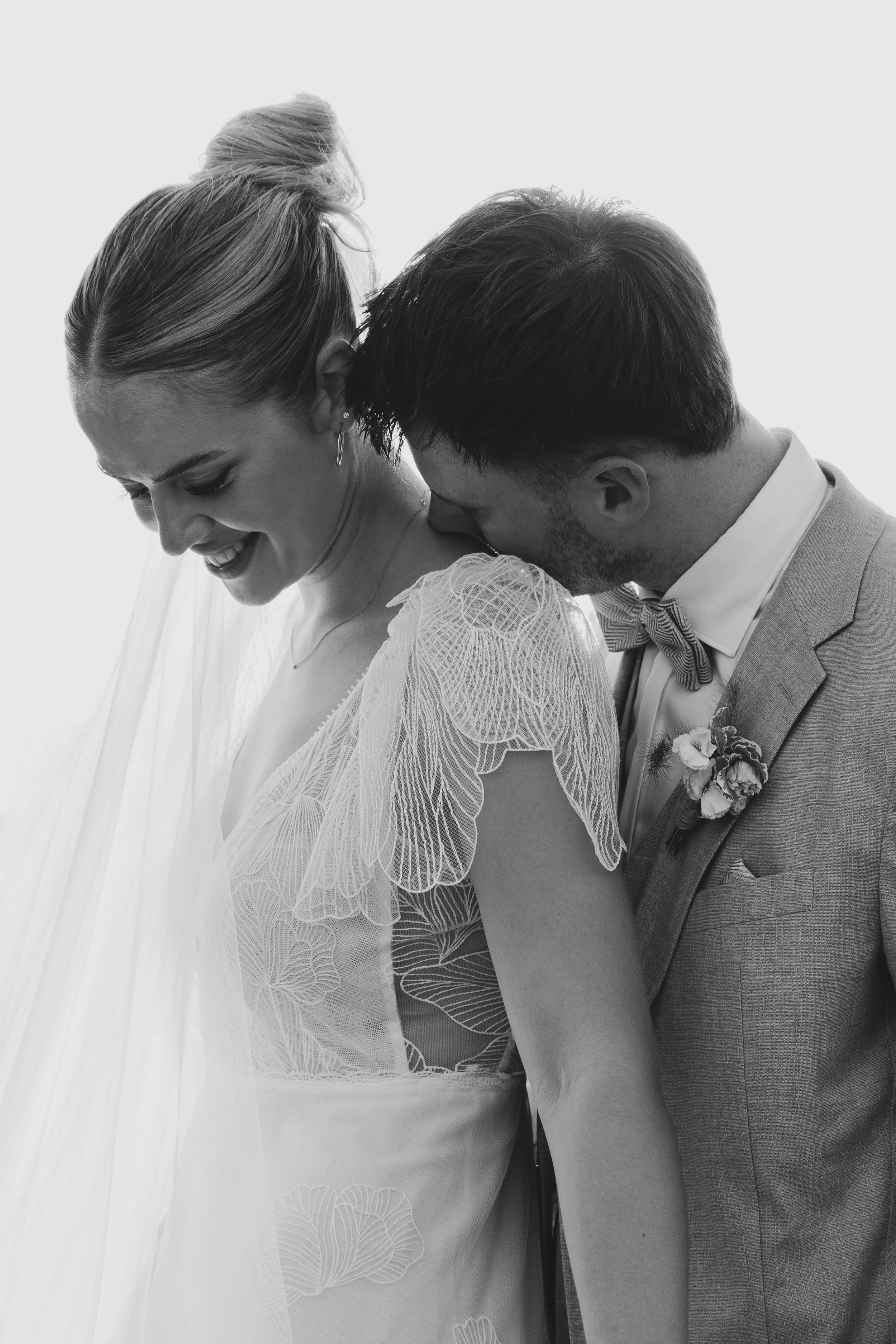 Ein Hochzeitsfoto eines Paares in formeller Kleidung, wobei der Mann der Frau liebevoll am Hals und Gesicht küsst, beide lächelnd und glücklich.