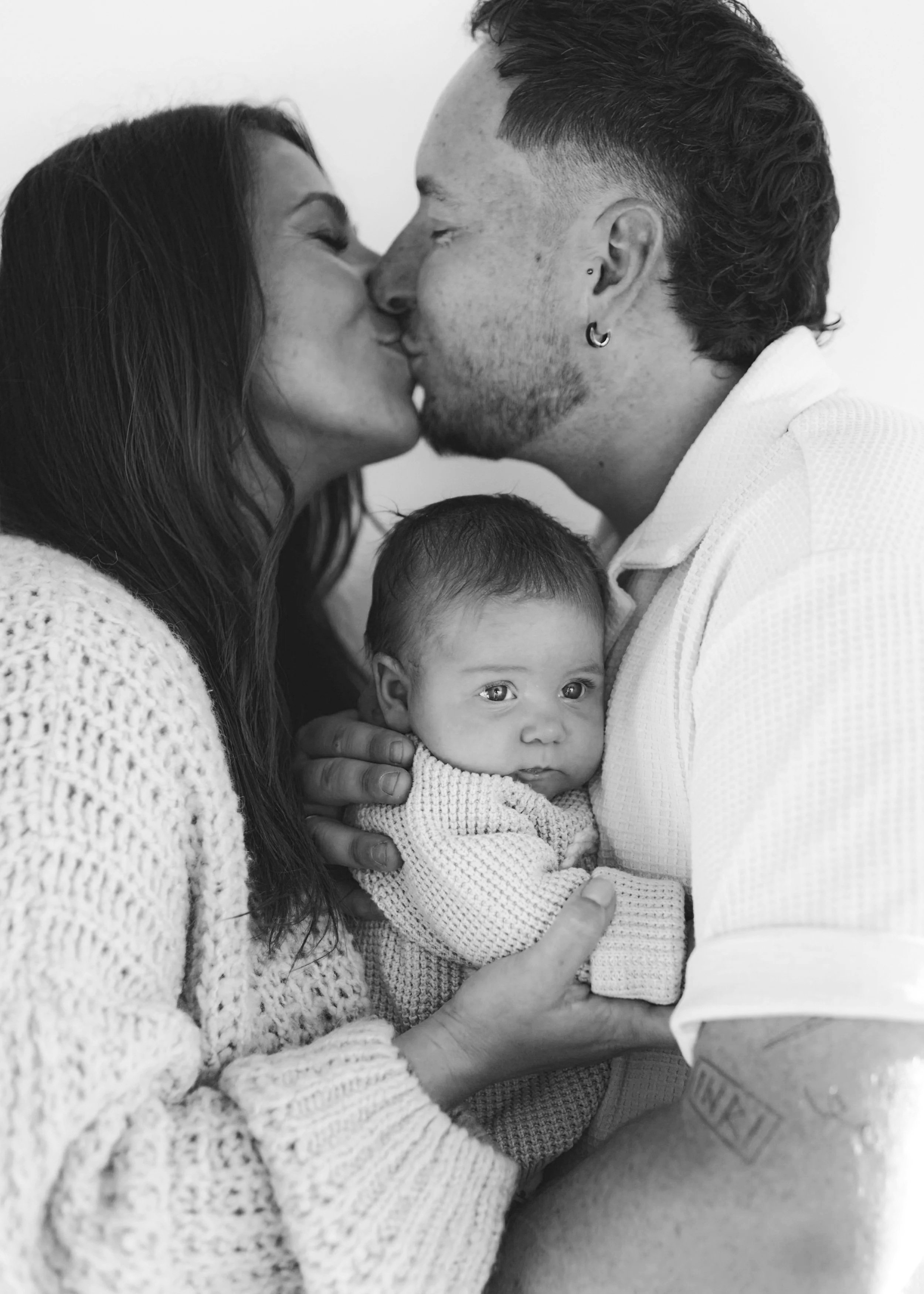 Ein schwarz-weiß Foto einer Familie: ein Paar küsst sich, während sie ein Baby halten. Das Baby schaut direkt in die Kamera.