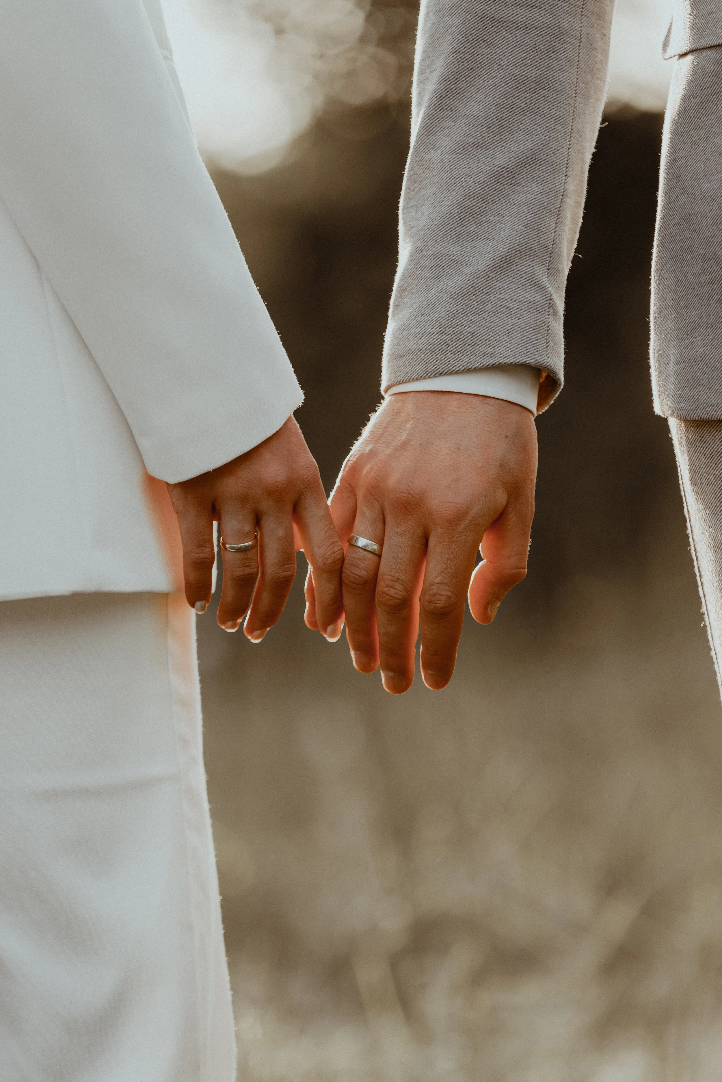 Nahaufnahme von zwei Paar Händen, die sich berühren, mit Eheringen an den Fingern, im Hintergrund sind die Unterkörper zweier Menschen in formeller Kleidung bei Sonnenuntergang.