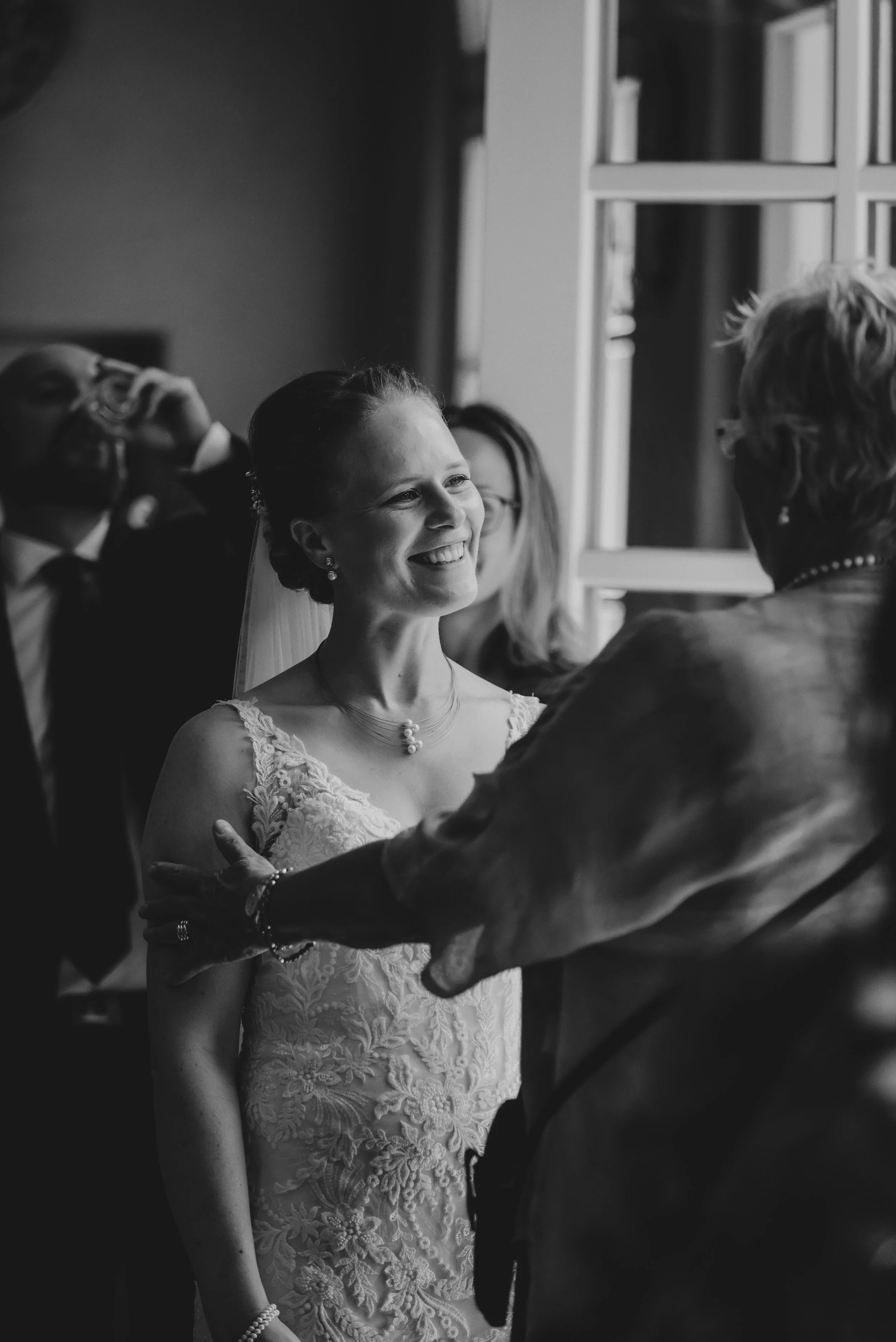 Eine glückliche Braut lächelt während ihrer Hochzeitszeremonie, umgeben von Angehörigen, in einem Raum mit großem Fenster.