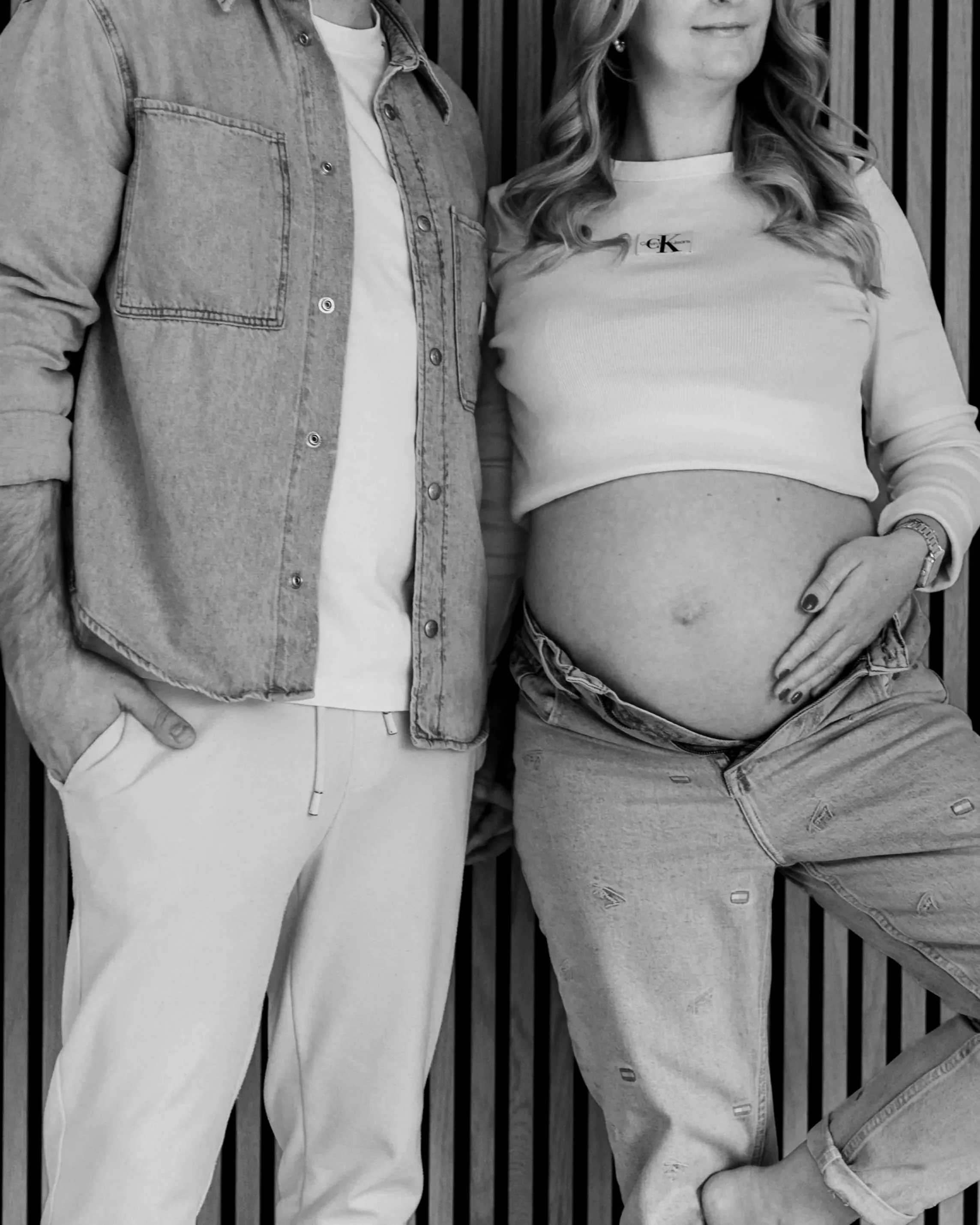 Schwangere Frau und Mann stehen nebeneinander vor einer Holzverkleidung, die Frau zeigt ihren Babybauch, er trägt leger Kleidung.