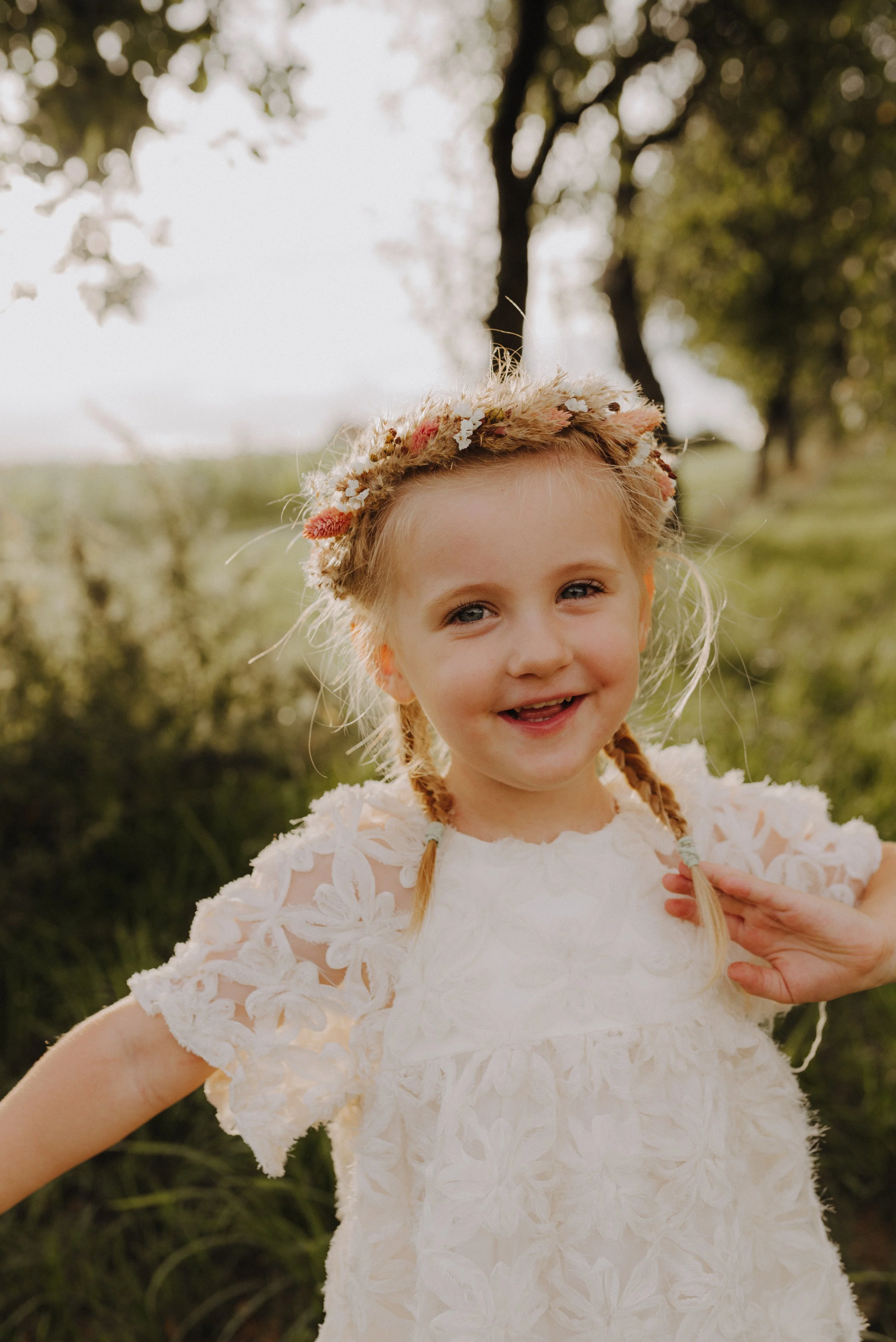 Ein lachendes Mädchen mit Zöpfen, trägt eine weiße geblümte Schleierrock, im Freien unter Bäumen im Sonnenlicht, mit Blumenkranz im Haar.