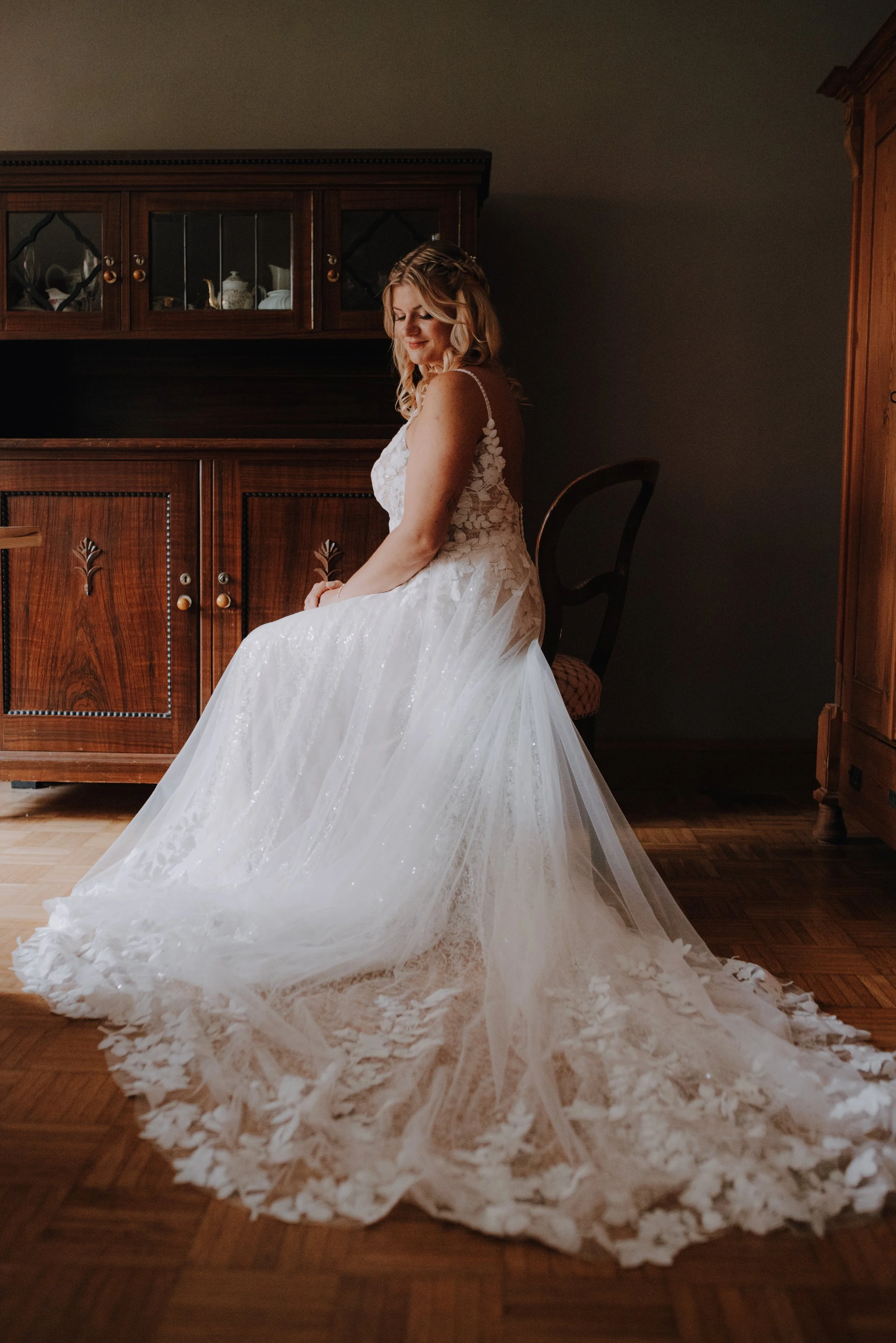 Eine Braut in einem weißen Hochzeitskleid sitzt in einem Raum mit Holzmöbeln.