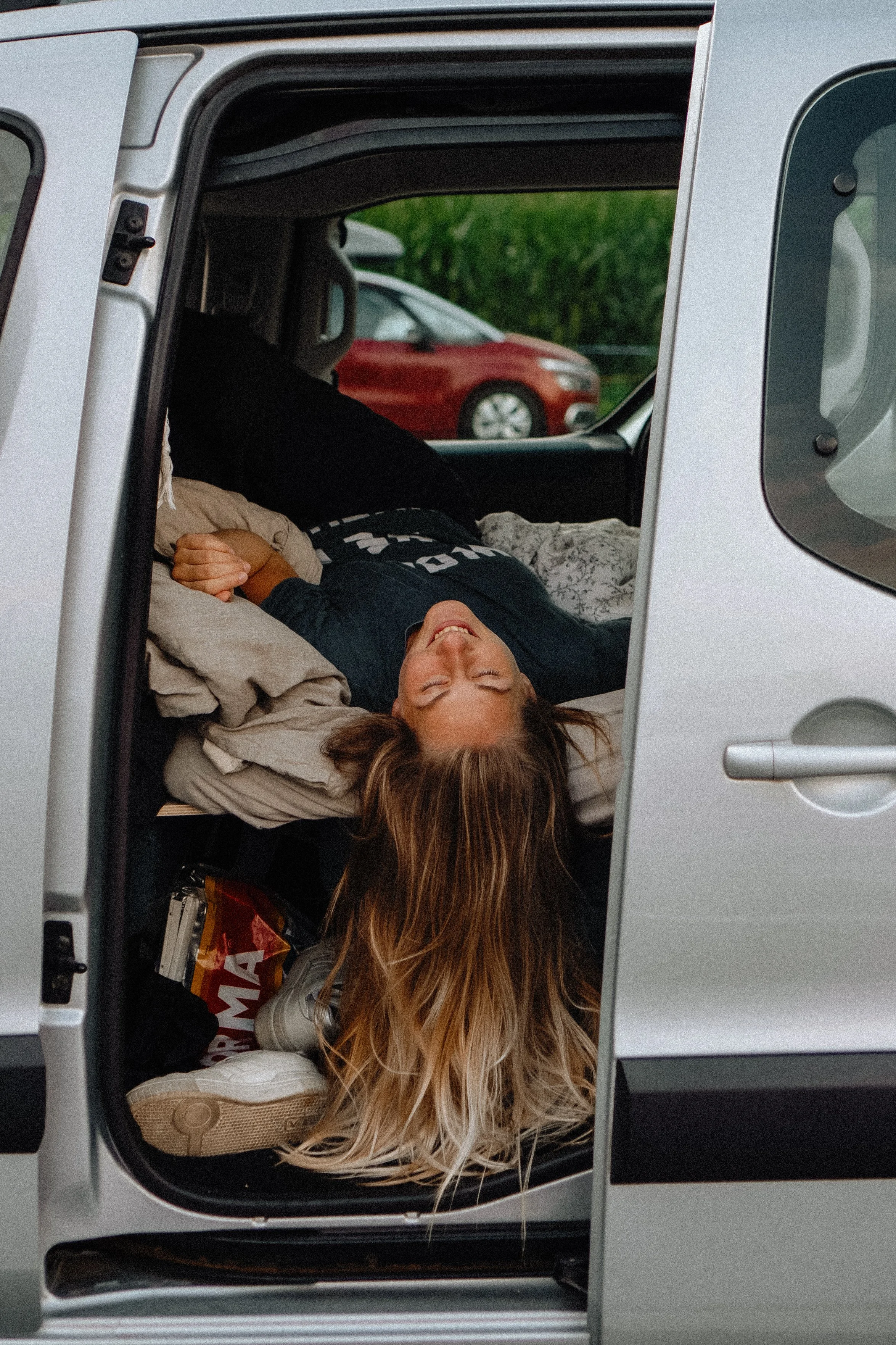 Frau liegt im Auto im Hinterraum und schläft mit Lächeln, umgeben von Gepäck und Gegenständen.