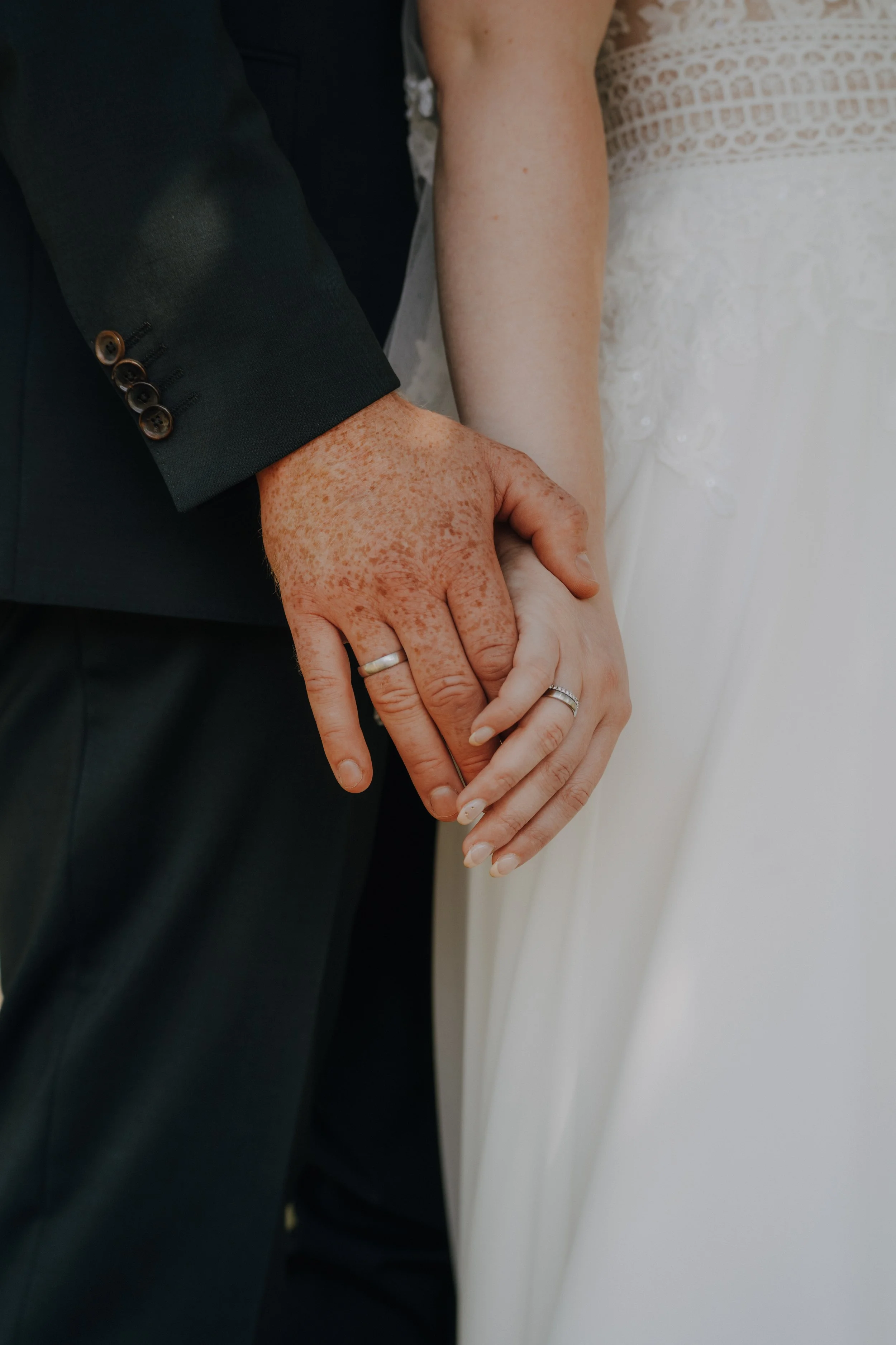 Nahaufnahme der Hände eines Paares an ihrer Hochzeitsfeier: Ein Mann in einem schwarzen Anzug mit einem Ehering auf dem Ringfinger hält die Hand einer Frau in einem weißen Brautkleid, die ebenfalls einen Ehering trägt.