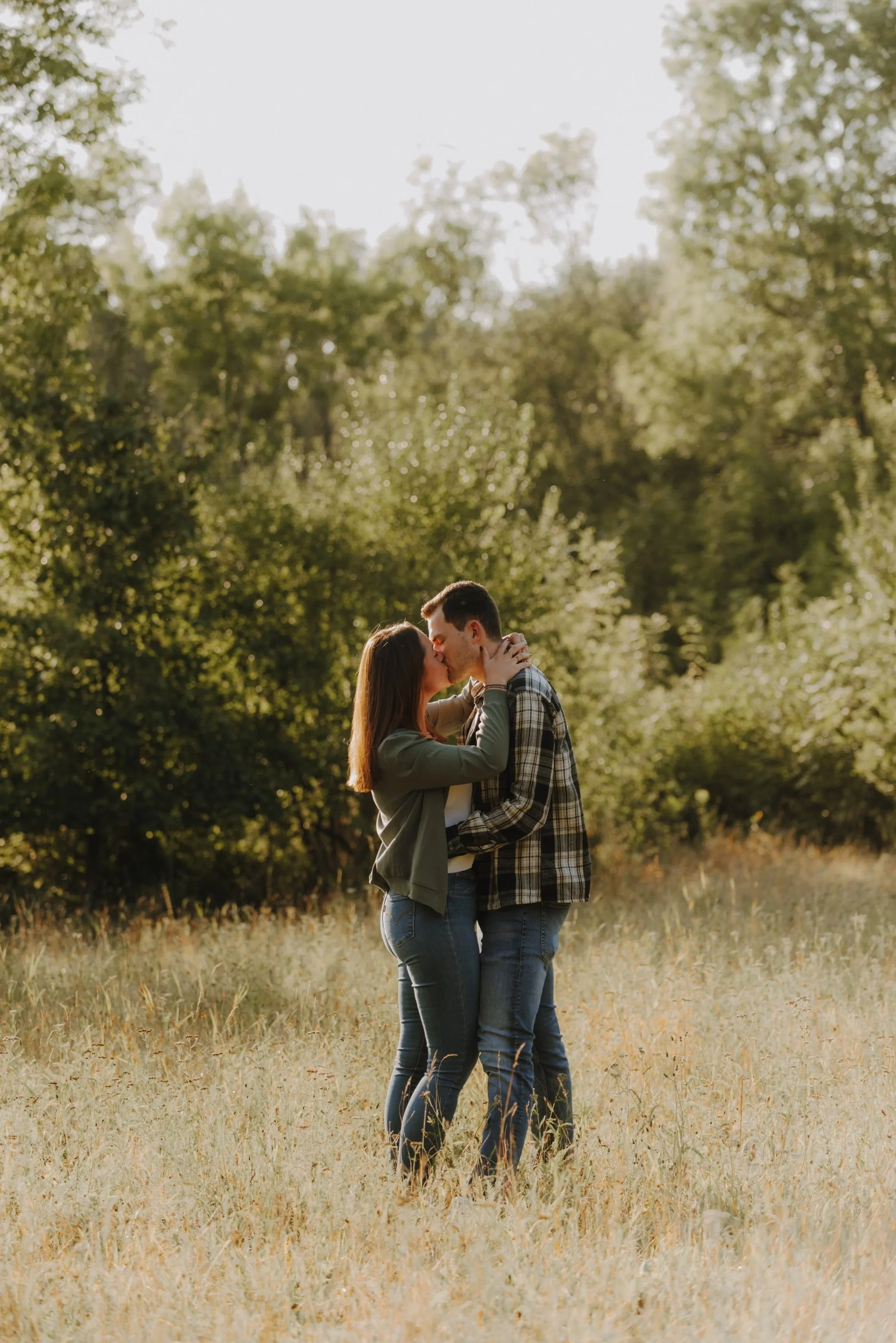 Ein junges Paar küsst sich liebevoll im Freien, umgeben von Bäumen und Gras. Es ist wahrscheinlich Sonnenuntergang oder früher Abend, was der Szene einen warmen Ton verleiht.