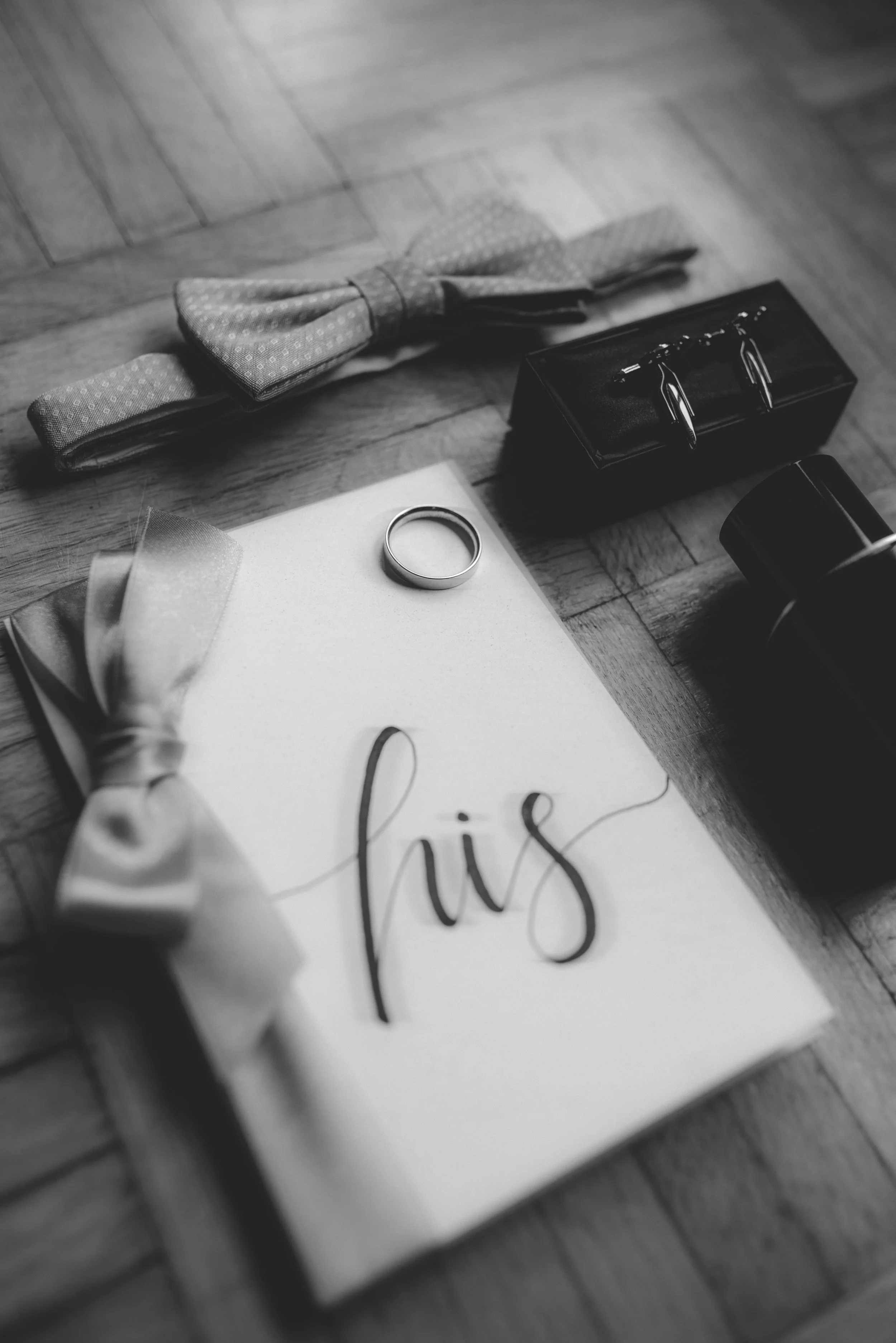 Ein weißes Gästebuch mit der Aufschrift 'his' in eleganter Schrift, ein Ehering, eine Stoffschleife, eine Flasche Parfüm, eine Krawatte, ein Koffer und ein Hochzeitssymbol liegen auf einem Holzboden.