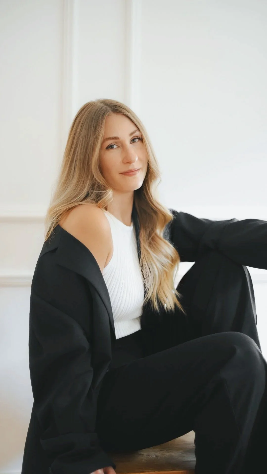 Junge Frau mit blonden, langen, gewellten Haaren sitzt auf dem Boden in einem weißen Raum, trägt eine weiße Bluse und eine schwarze Jacke, schaut lächelnd in die Kamera.