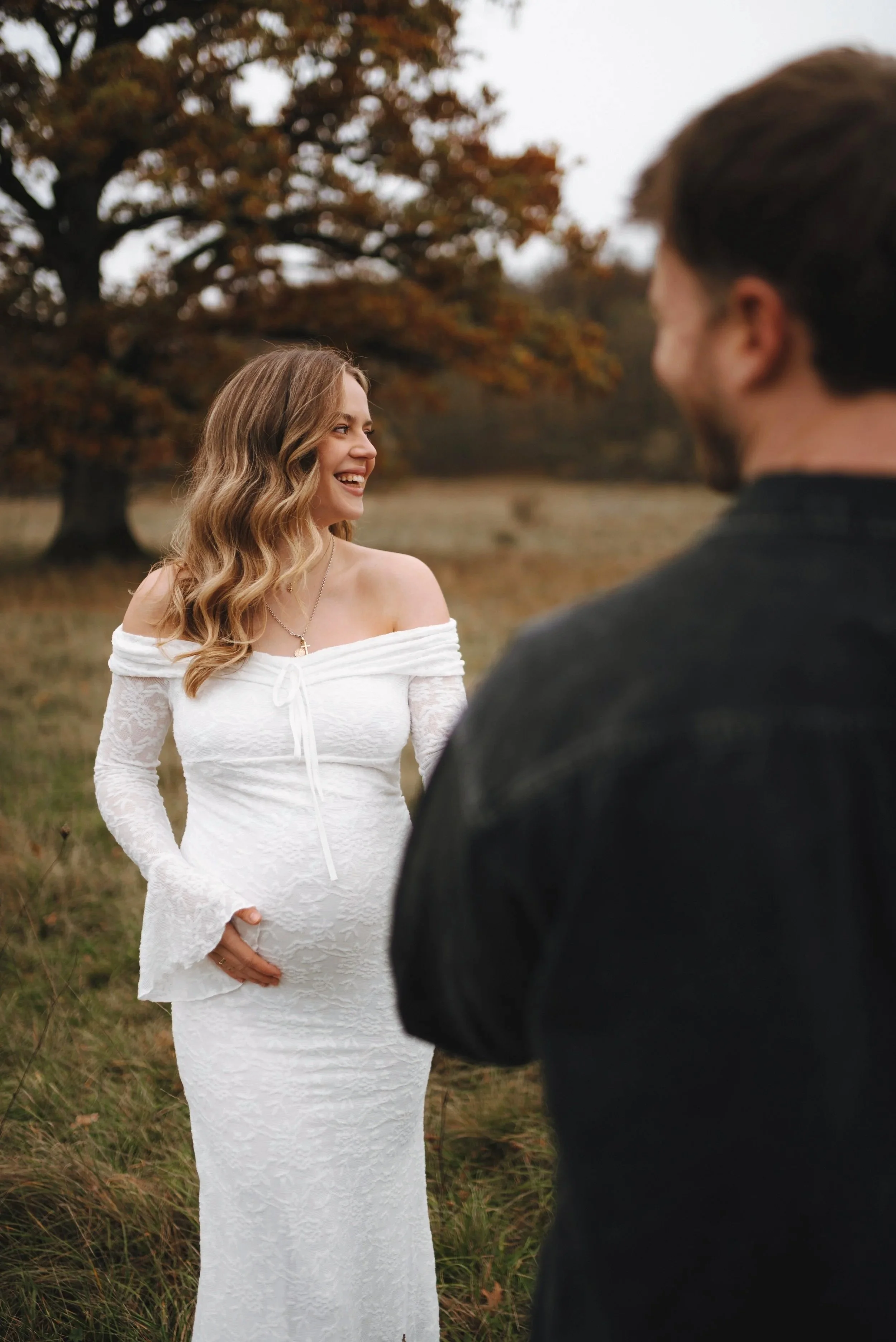 Ein lächelndes Brautpaar steht draußen in einem Feld mit herbstlichen Bäumen im Hintergrund, die Braut trägt ein schulterfreies, langärmeliges weißes Brautkleid, der Bräutigam trägt schwarzen Hemd.