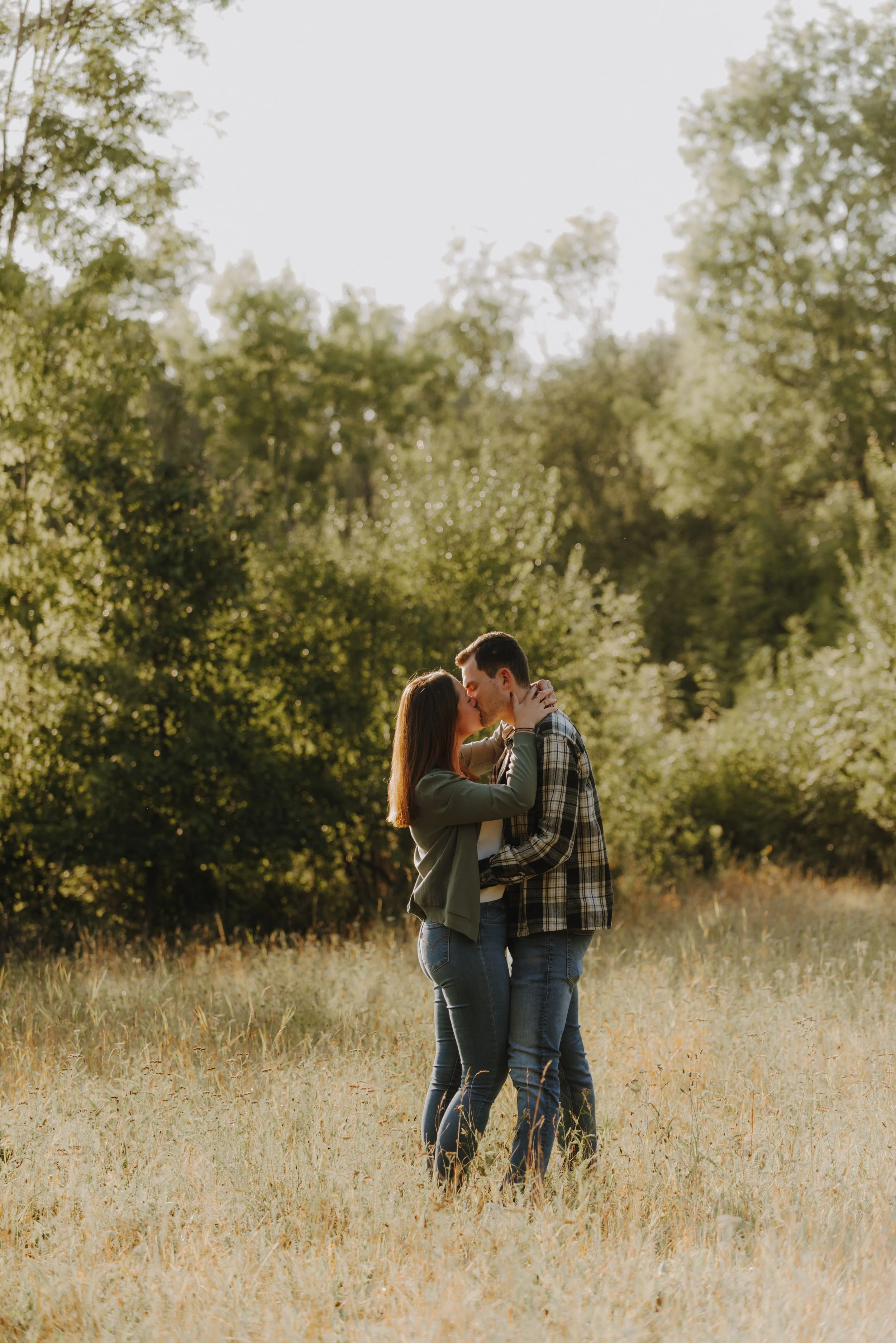 Ein verliebtes Paar umarmt sich und küsst sich auf einer Wiese im Freien, umgeben von Bäumen, bei Sonnenuntergang.
